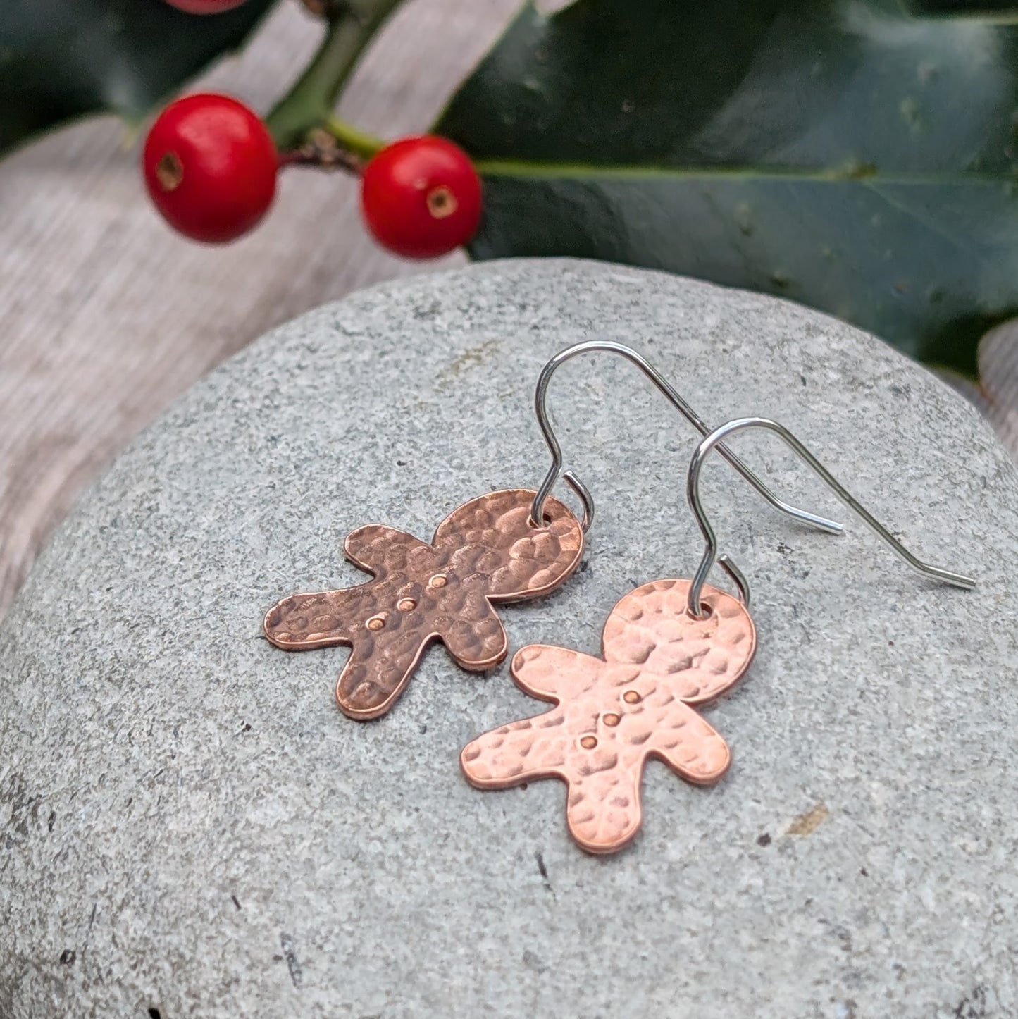 Side view of a pair of handmade copper gingerbread drop earrings with a hammered finish, resting on a grey stone surface, with holly berries in the background.