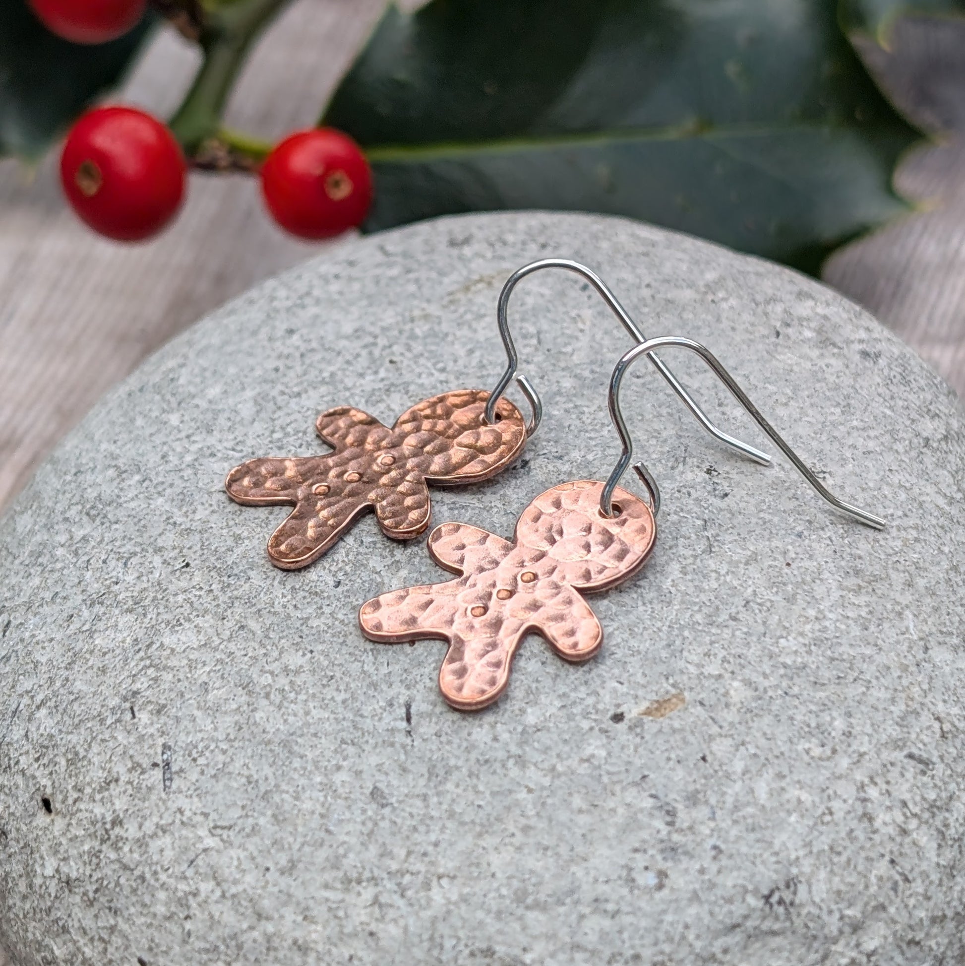 Side view of a pair of handmade copper gingerbread drop earrings with a hammered finish, resting on a grey stone surface, with holly berries in the background.