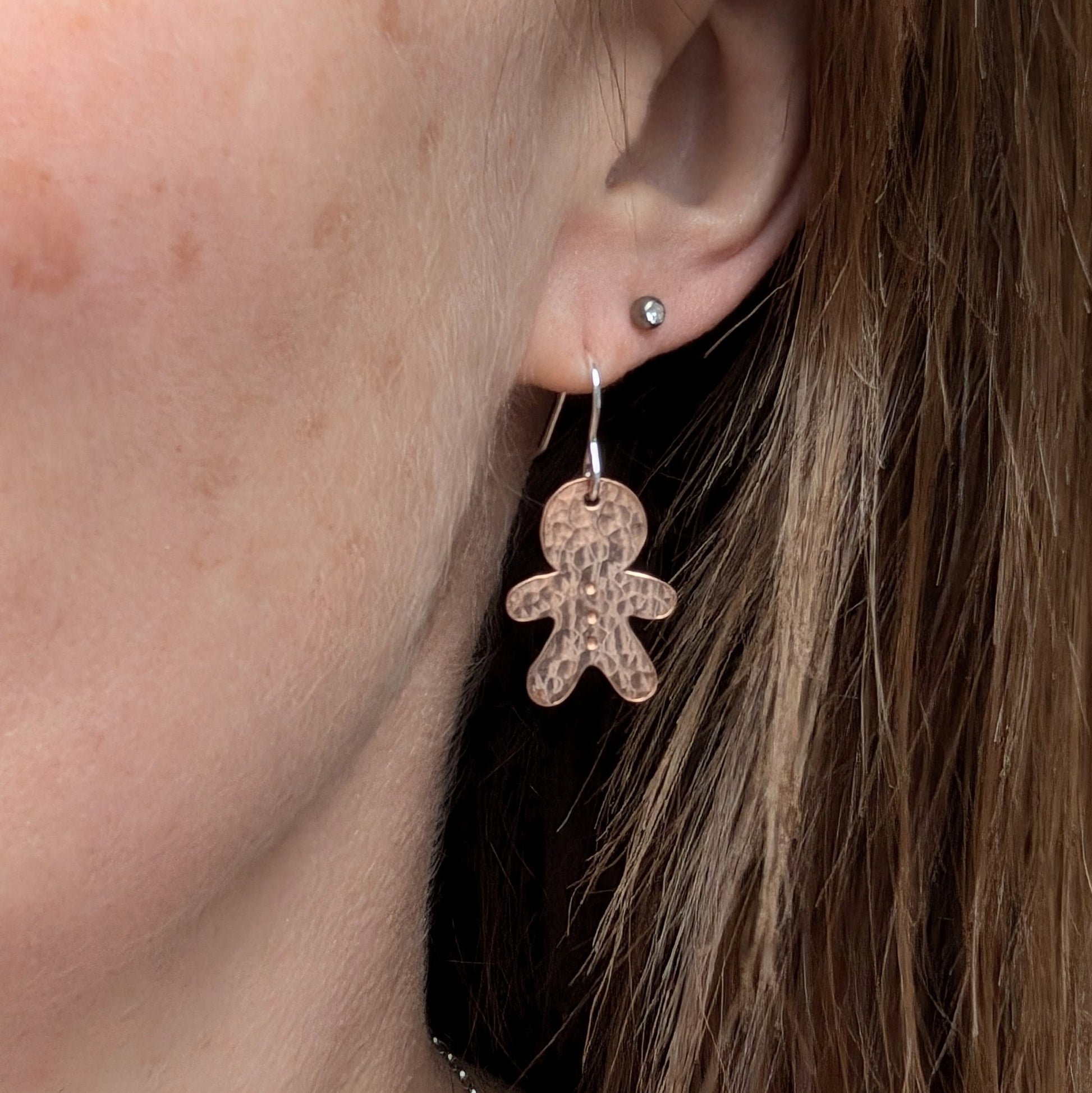 Close-up of a copper gingerbread drop earring being worn, showing the hammered texture and sterling silver ear wire, with another stud earring visible in a second piercing.