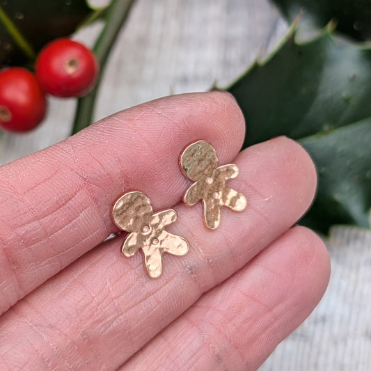 Close-up of a pair of handmade copper gingerbread stud earrings with a hammered texture, being held in a person's hand, with a festive holly branch and berries blurred in the background.