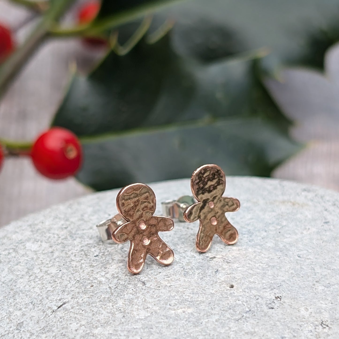 Close-up of handmade copper gingerbread stud earrings with a hammered finish, displayed on a grey stone surface with festive holly and berries in the background.