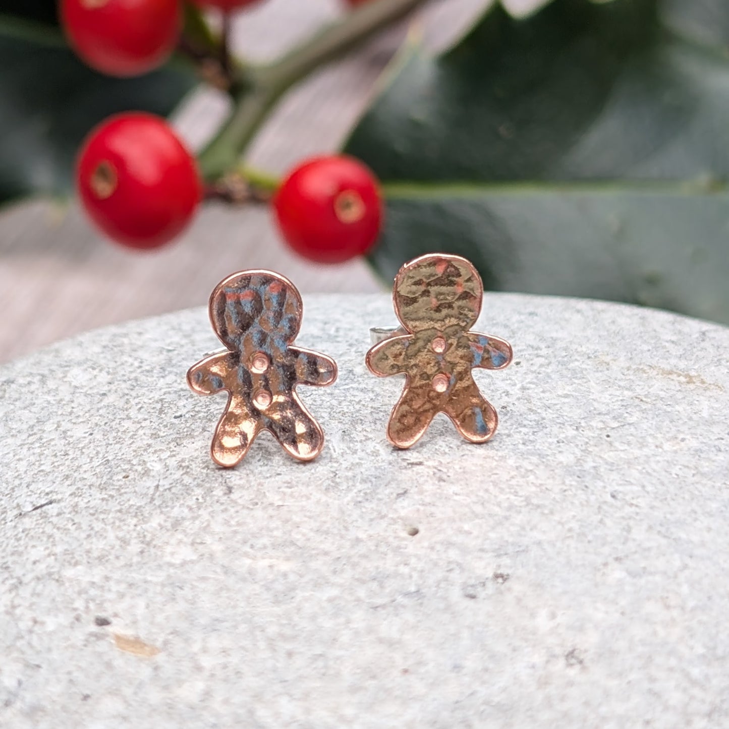 Close-up of handmade copper gingerbread stud earrings with a hammered finish, displayed on a grey stone surface with festive holly and berries in the background.