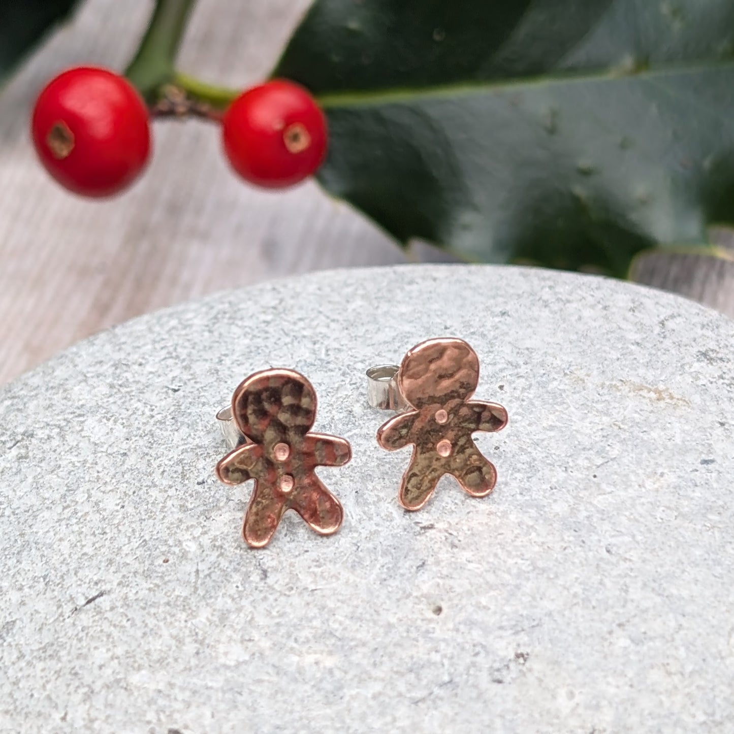 Close-up of handmade copper gingerbread stud earrings with a hammered finish, displayed on a grey stone surface with festive holly and berries in the background.