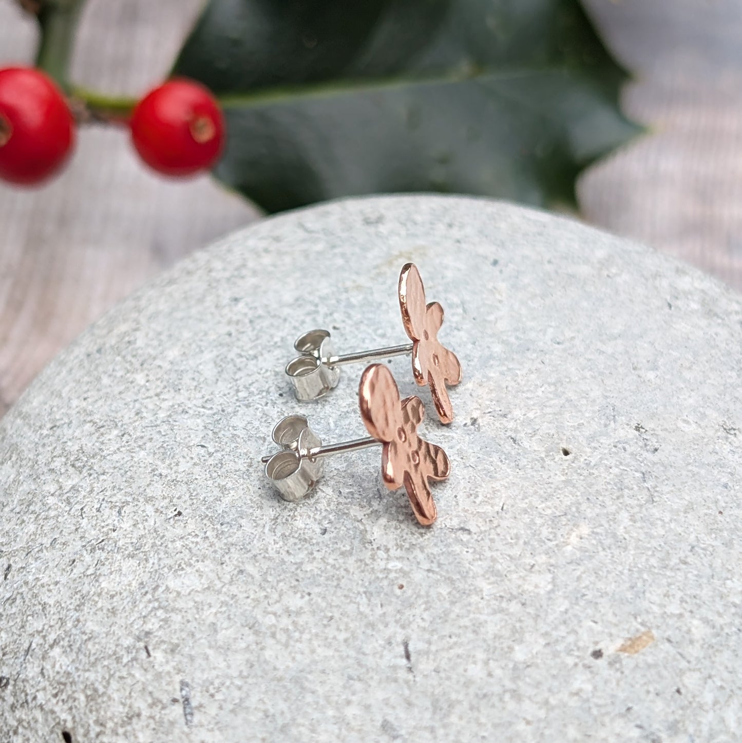 Side view of copper gingerbread stud earrings, showing their sterling silver posts, resting on a textured grey stone, with holly leaves and berries in the background.