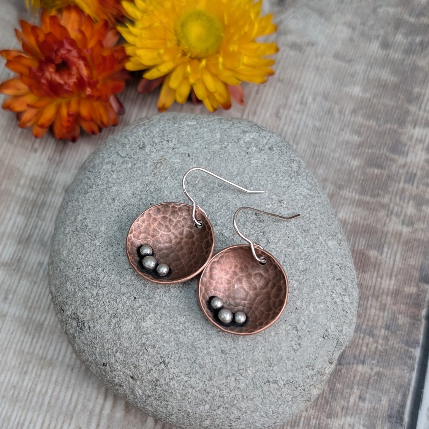 Each earring is an inverted copper hammered disc with three small silver pebbles on the lower curve of the disc. The hammered texture is accentuated by the oxidised copper. Suspended from silver earring wire.