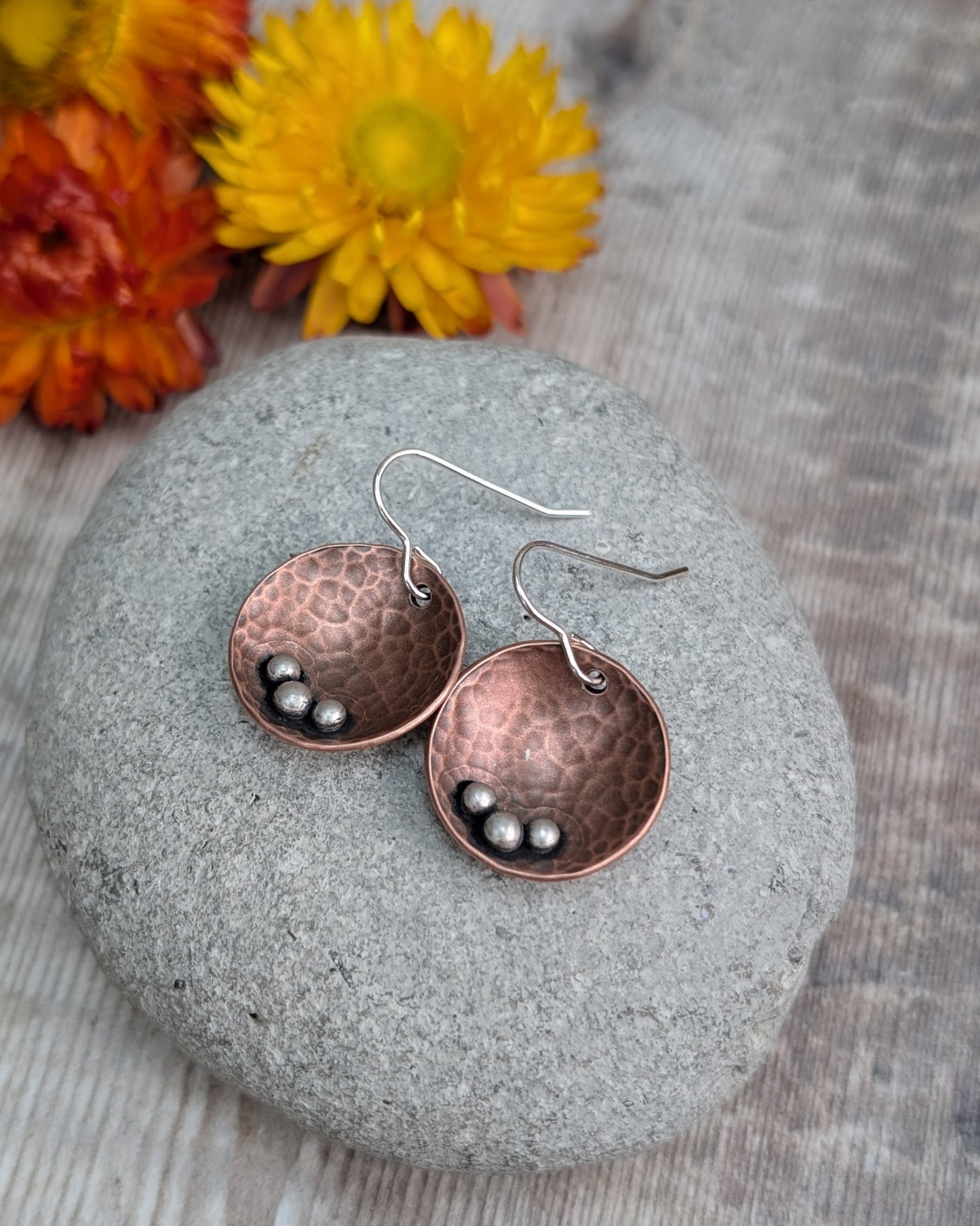 Each earring is an inverted copper hammered disc with three small silver pebbles on the lower curve of the disc. The hammered texture is accentuated by the oxidised copper. Suspended from silver earring wire.