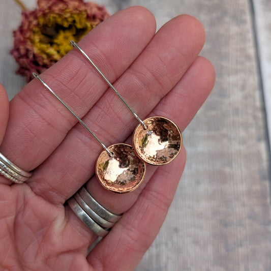 Close-up of a hand holding the hammered copper dome disc earrings by their long silver ear wires, showing the reflective, curved copper surface.