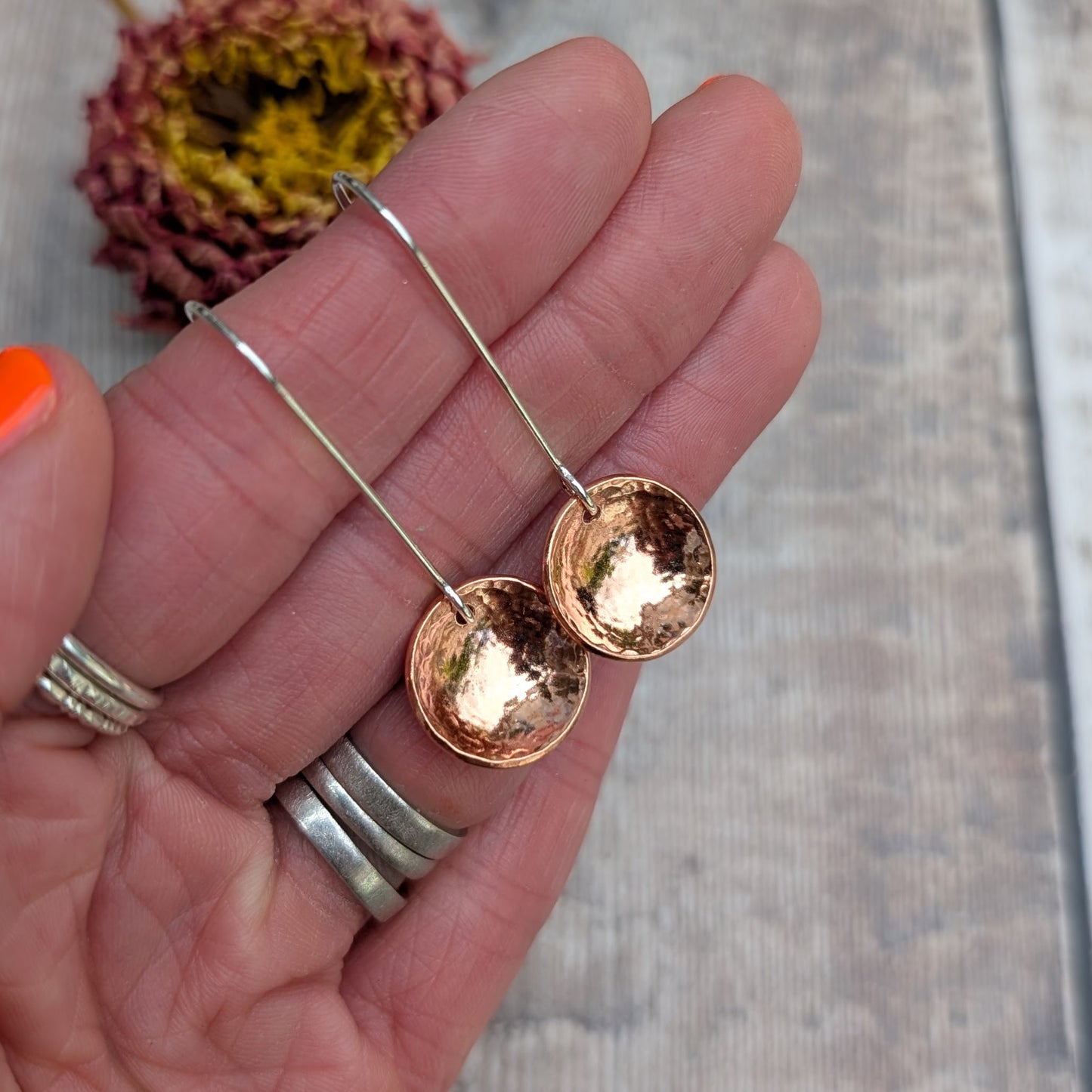 Close-up of a hand holding the hammered copper dome disc earrings by their long silver ear wires, showing the reflective, curved copper surface.