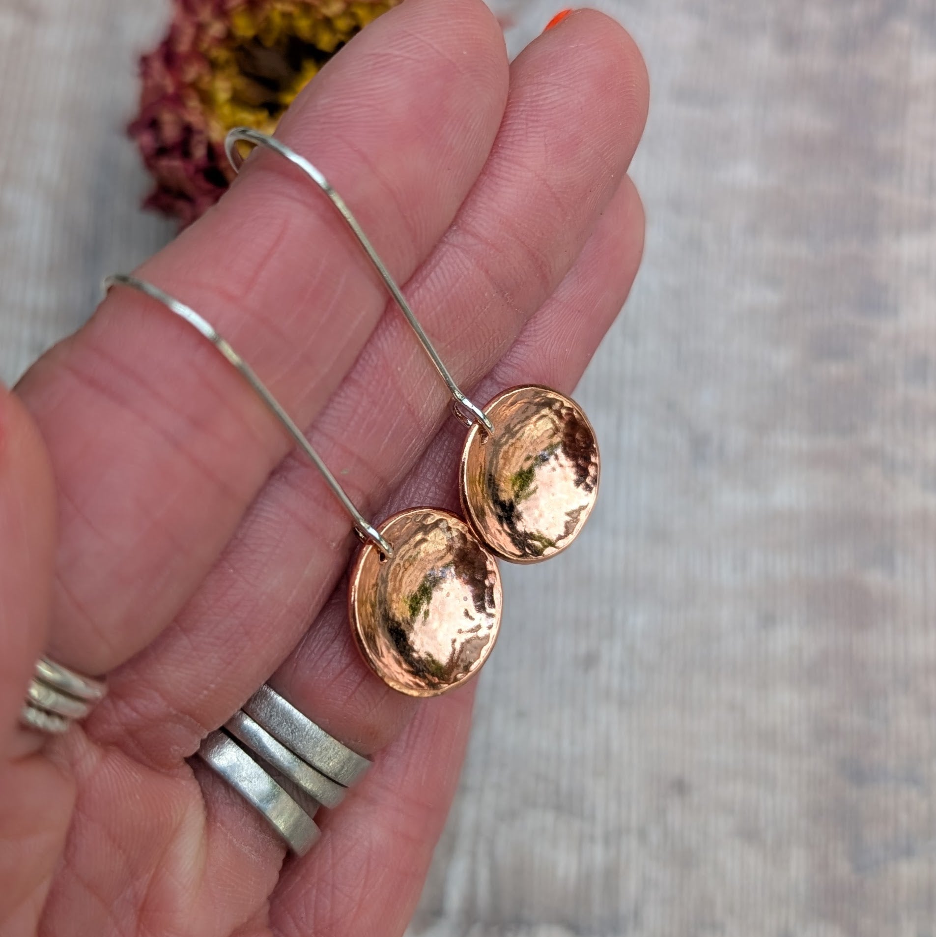 Close-up of a hand holding the hammered copper dome disc earrings by their long silver ear wires, showing the reflective, curved copper surface.