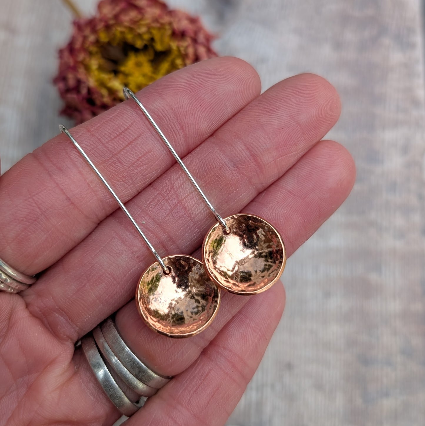 Close-up of a hand holding the hammered copper dome disc earrings by their long silver ear wires, showing the reflective, curved copper surface.