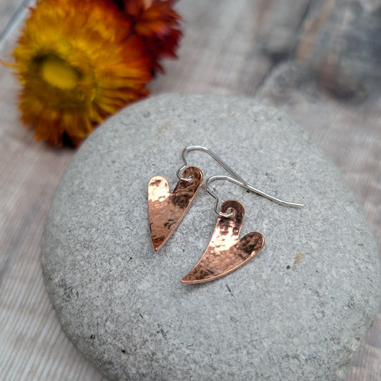 A pair of copper hammered heart earrings resting on a smooth grey stone, showing their gently curved shape and handmade texture, with an orange flower in the background.