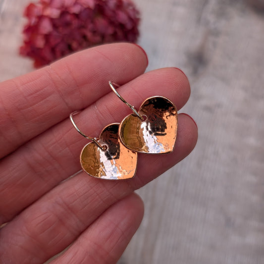 A close-up of hammered copper heart earrings held in a hand, showing their reflective surface.