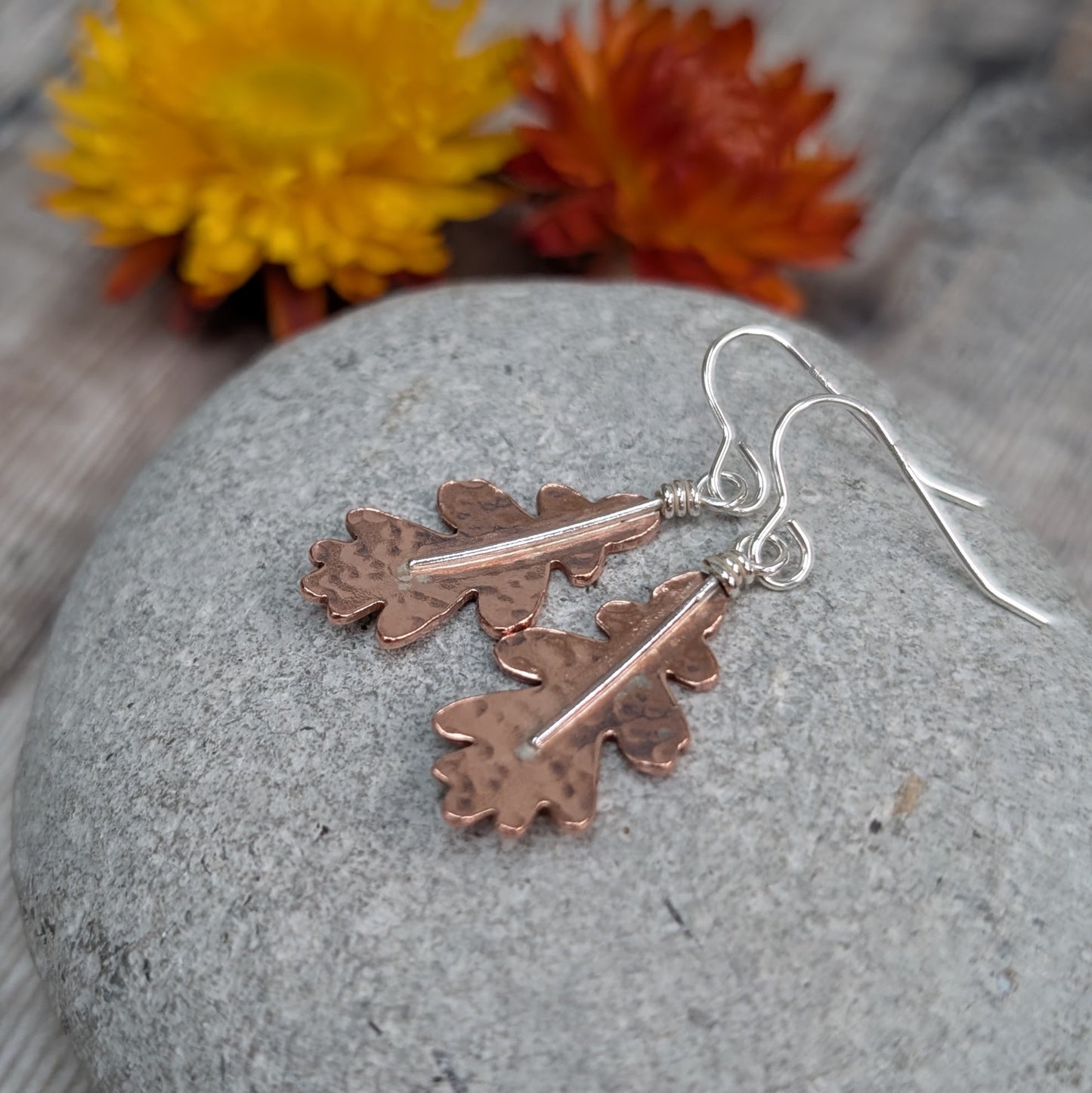 Copper hammered oak leaf earrings resting on a smooth grey stone, with bright orange and yellow dried flowers softly blurred in the background.