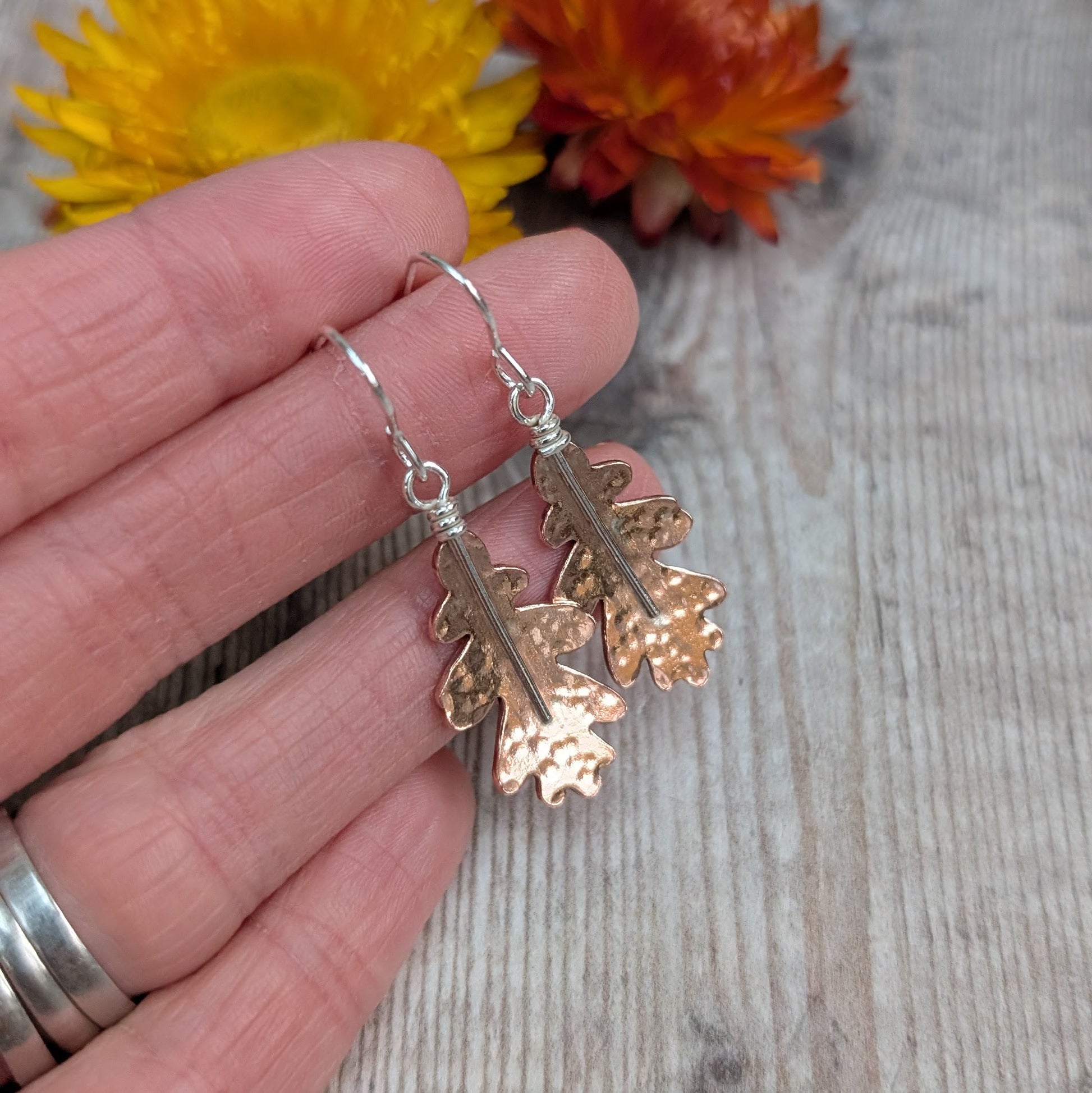 A hand holding a pair of copper hammered oak leaf earrings, showing their size and detail against a wooden background with dried flowers behind.