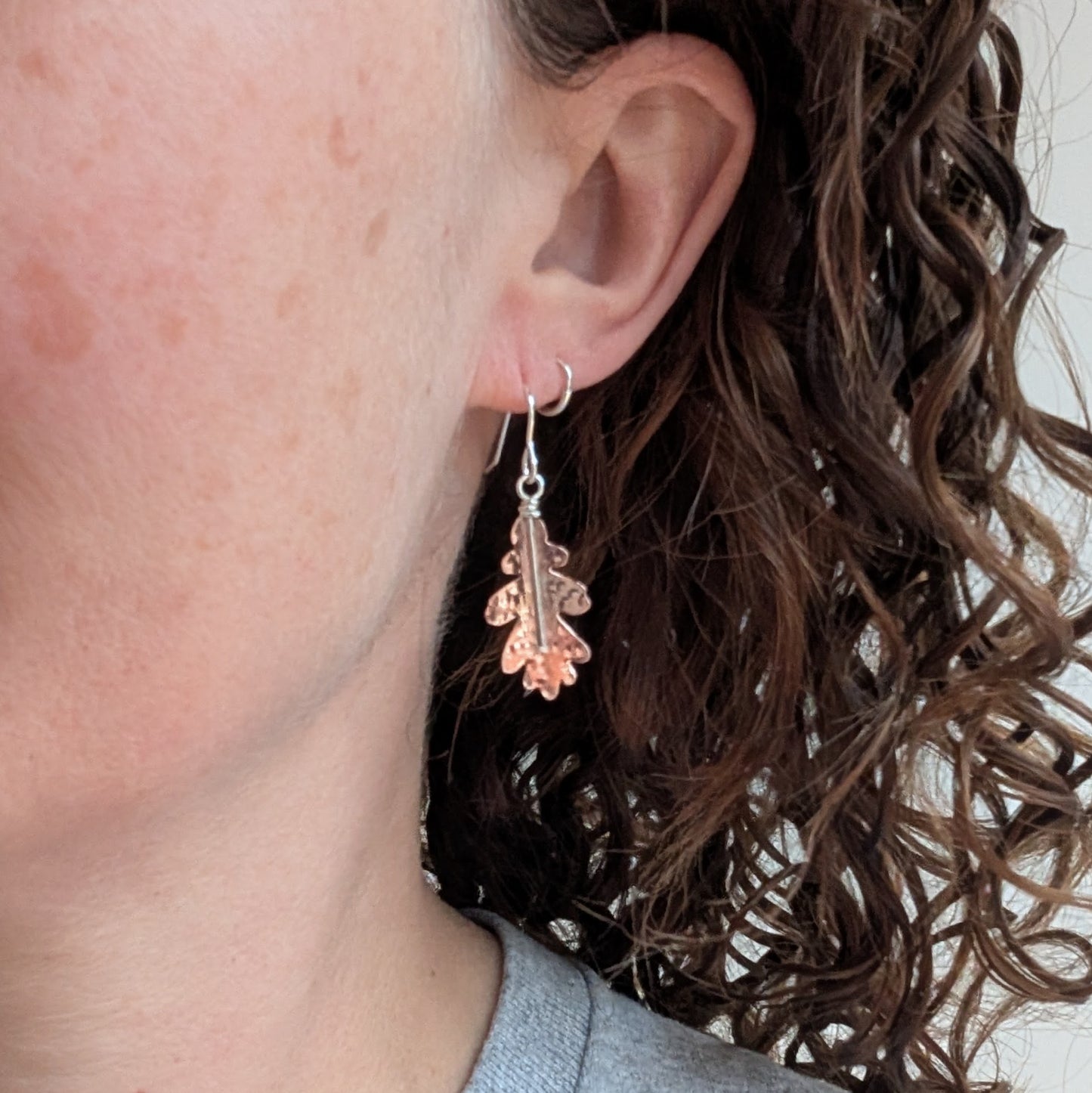 A close-up of a person wearing one copper hammered oak leaf earring, showing how it hangs and catches the light.