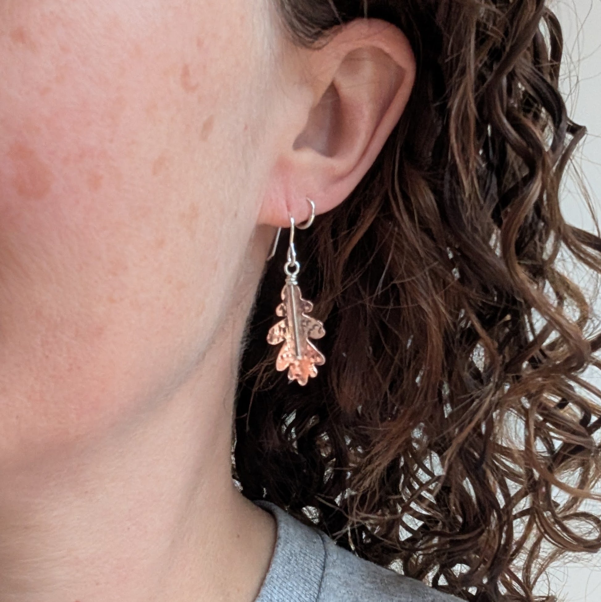 A close-up of a person wearing one copper hammered oak leaf earring, showing how it hangs and catches the light.