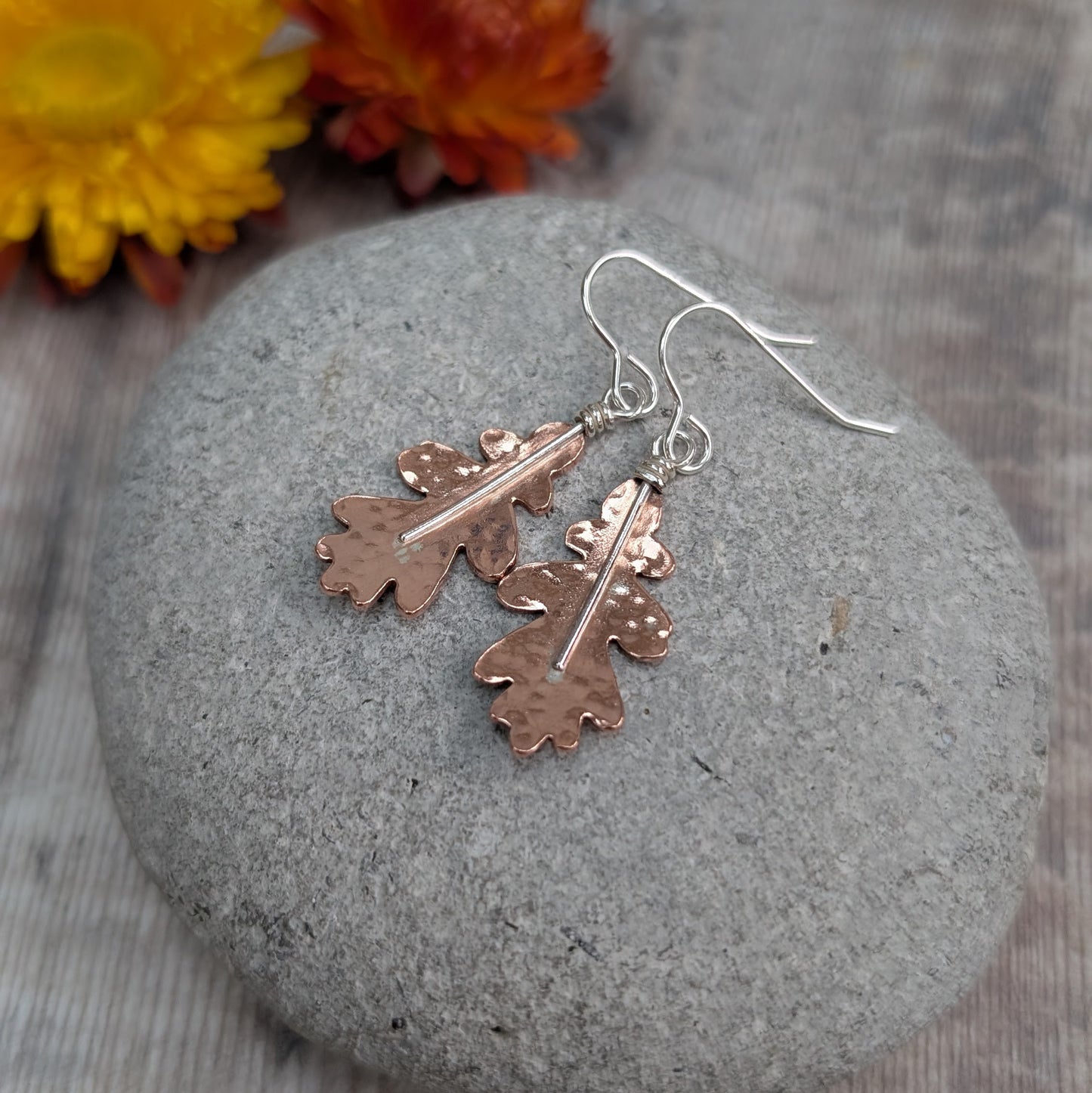 Copper hammered oak leaf earrings displayed on a grey stone, photographed from above with warm-toned dried flowers in the background.