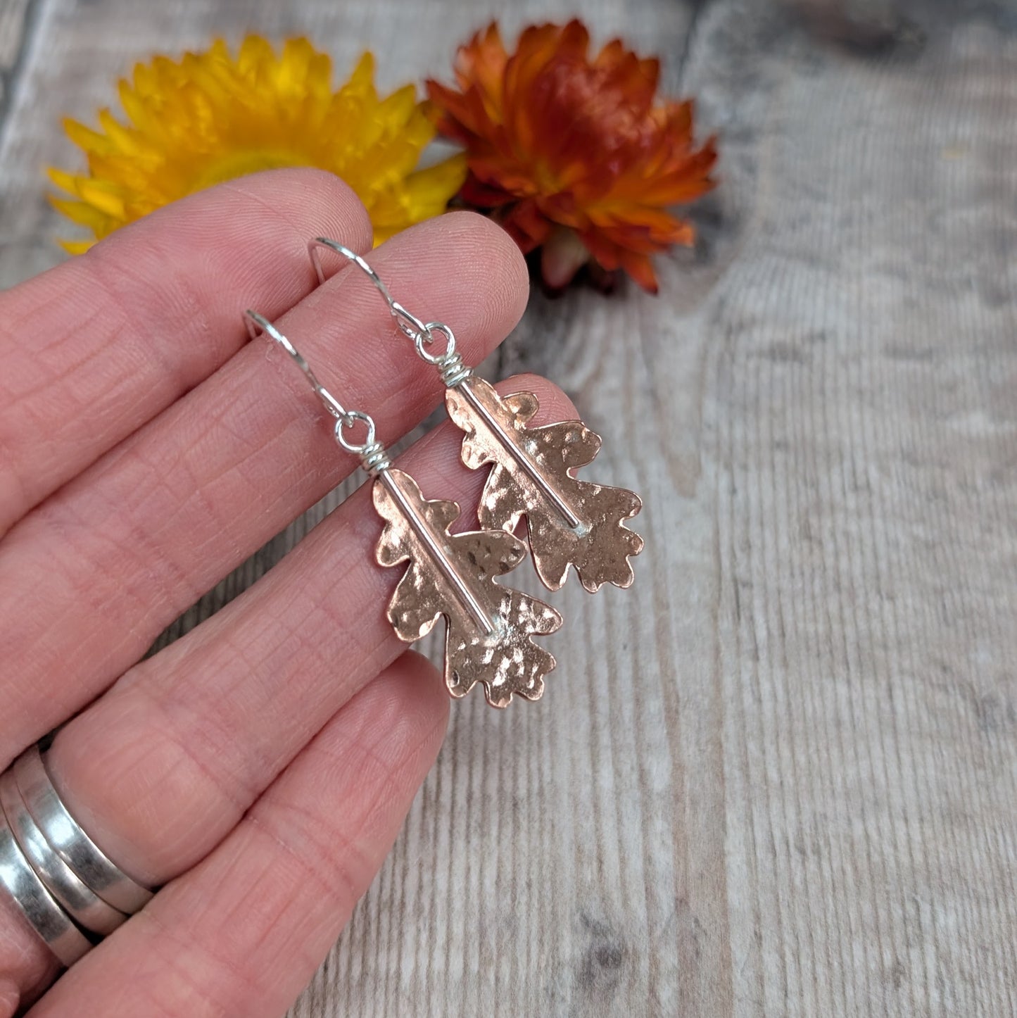 A hand holding the copper hammered oak leaf earrings, highlighting the gentle hammered finish on each leaf.