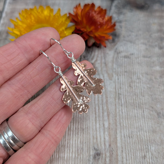 A hand holding the copper hammered oak leaf earrings, highlighting the gentle hammered finish on each leaf.
