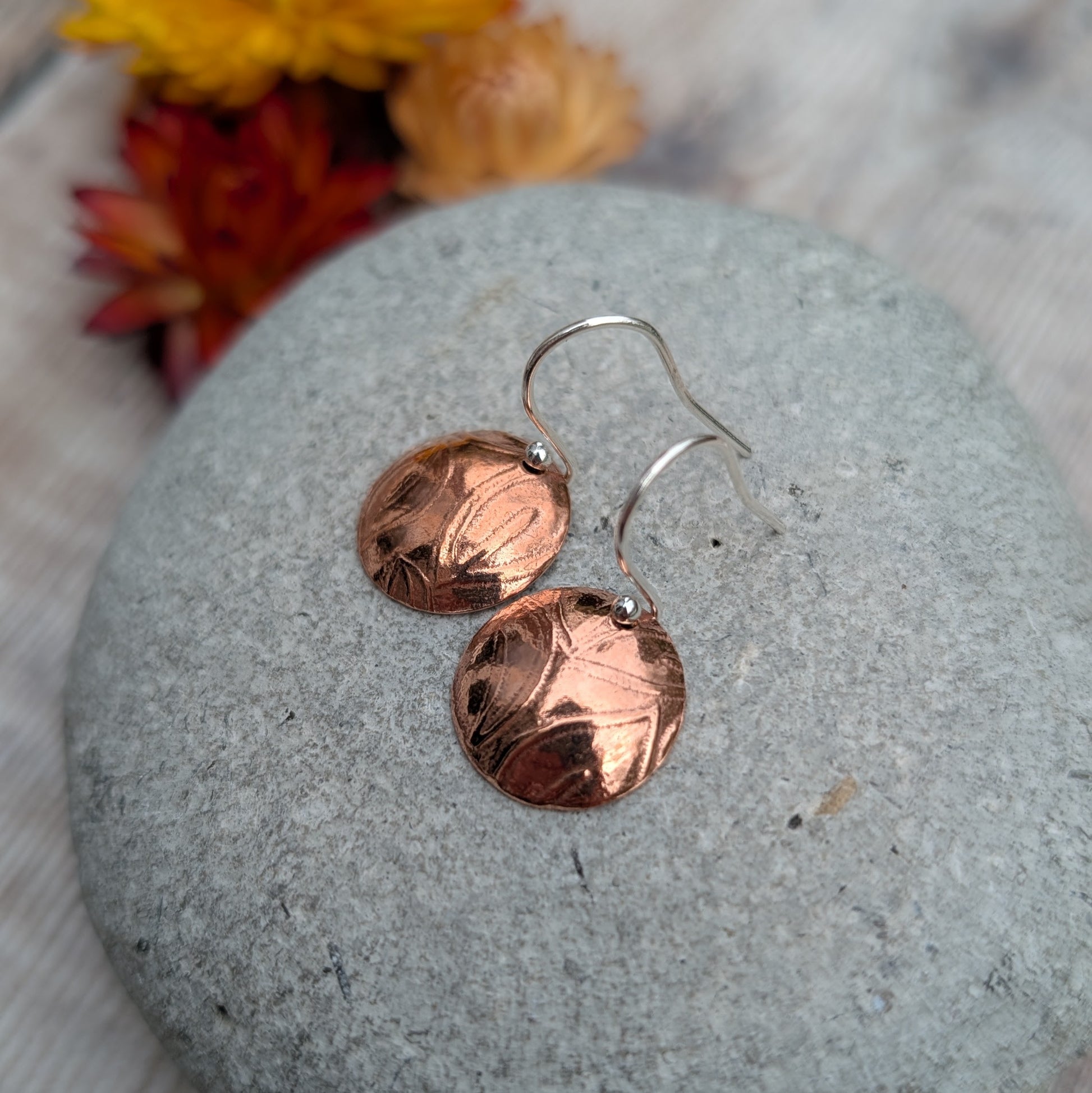Copper large leaf pattern dome disc earrings resting on a smooth grey stone showing the pressed bark-like leaf pattern.