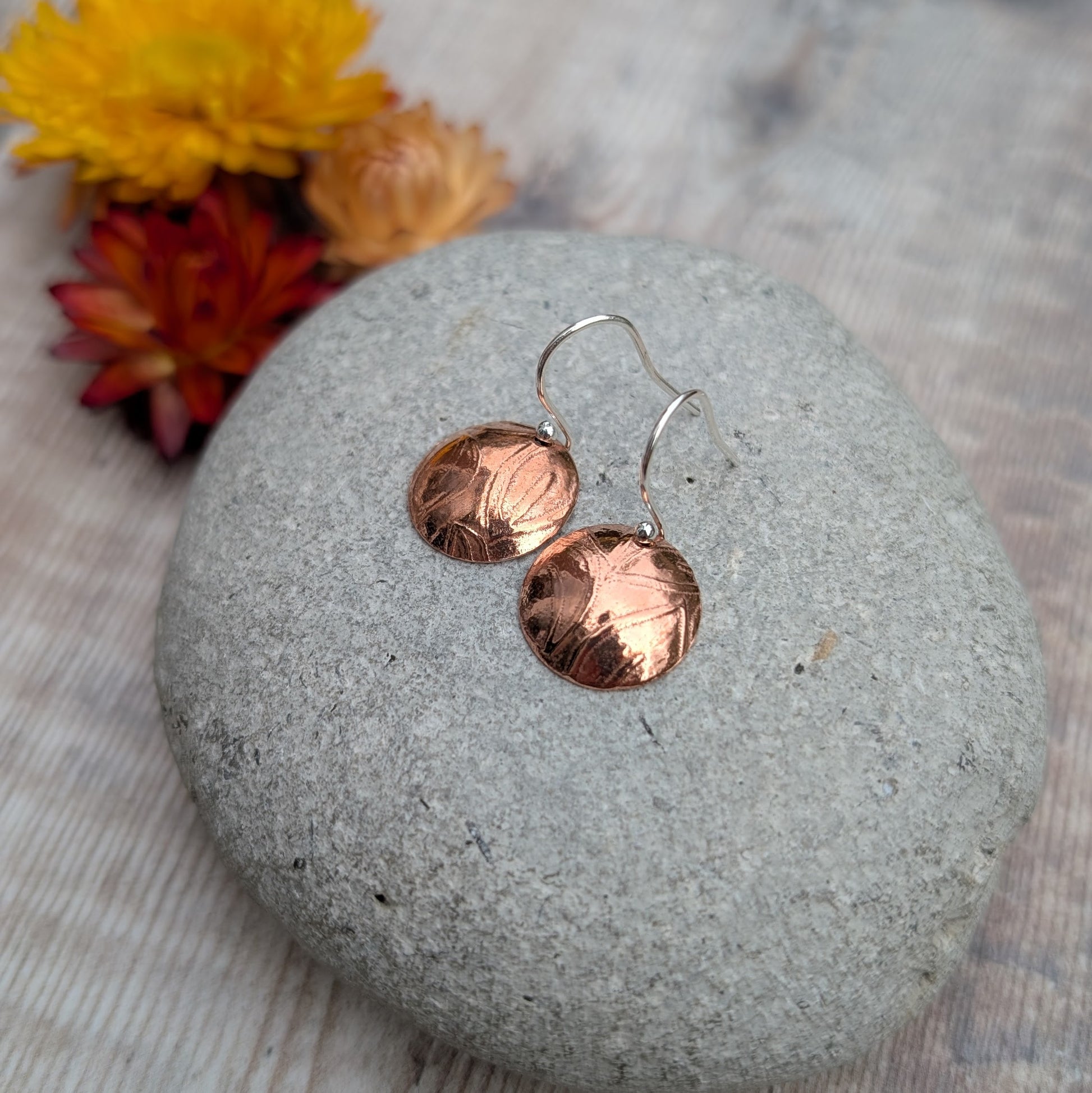 Pair of Copper large leaf pattern dome disc earrings displayed on a stone with dried flowers in the background.
