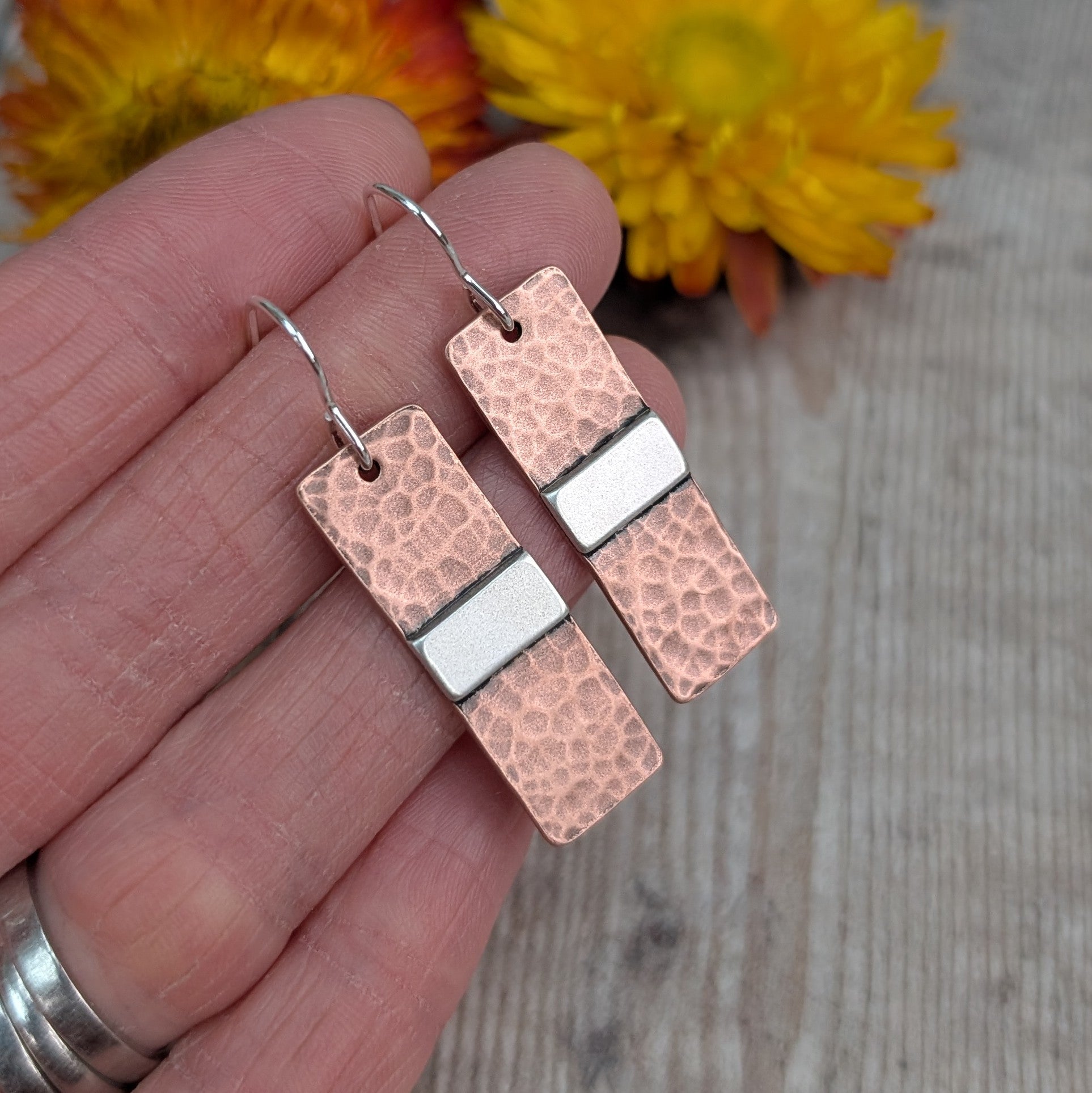 Each earring is a copper rectangle with a hammered oxidised finish. In the middle of the rectangle, there is a slightly raised silver strip. 