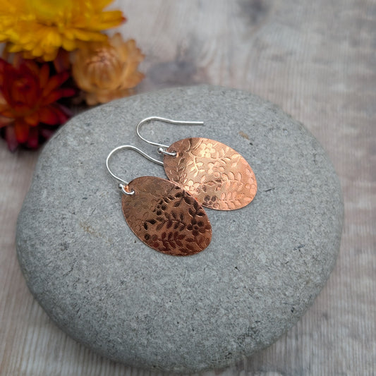 Copper floral pattern oval disc earrings resting on a smooth grey stone showing the pressed floral pattern.
