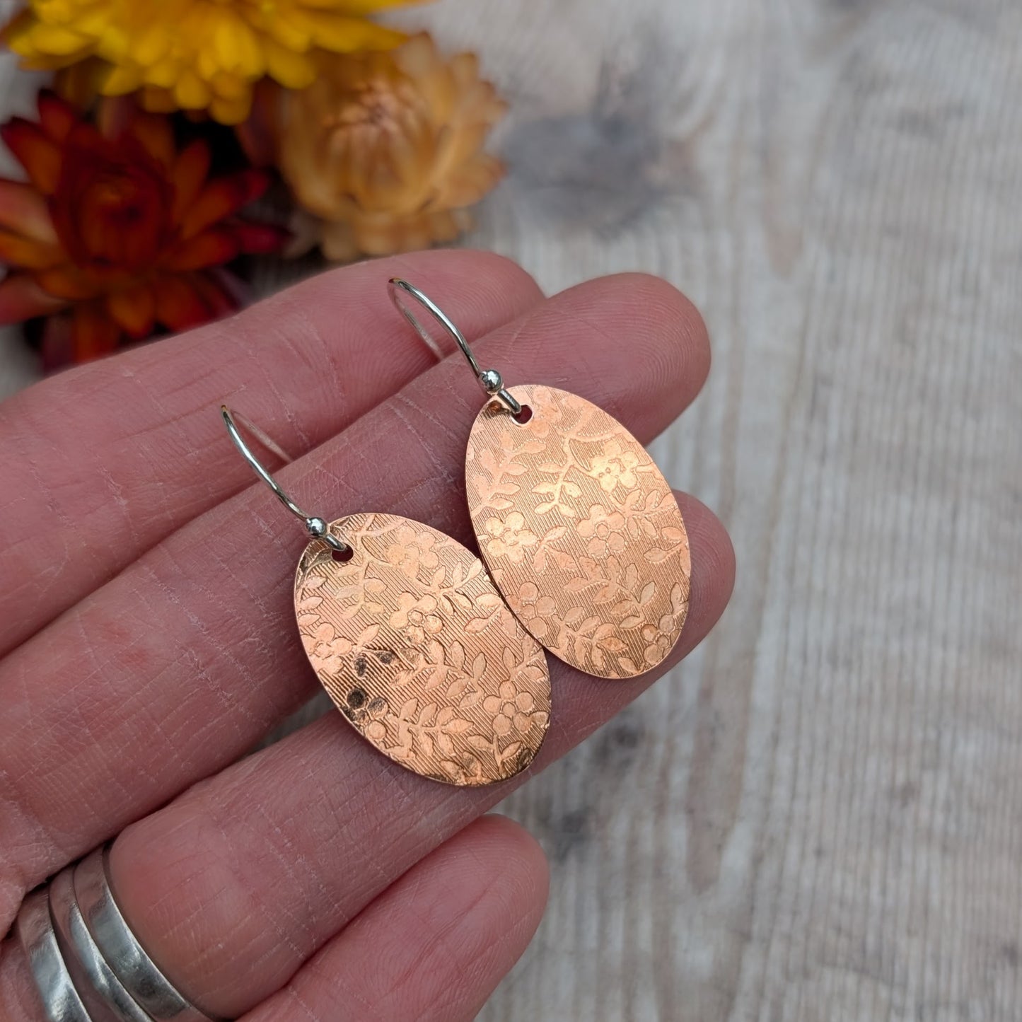 Copper floral pattern oval disc earrings held in a hand to show size and detail.