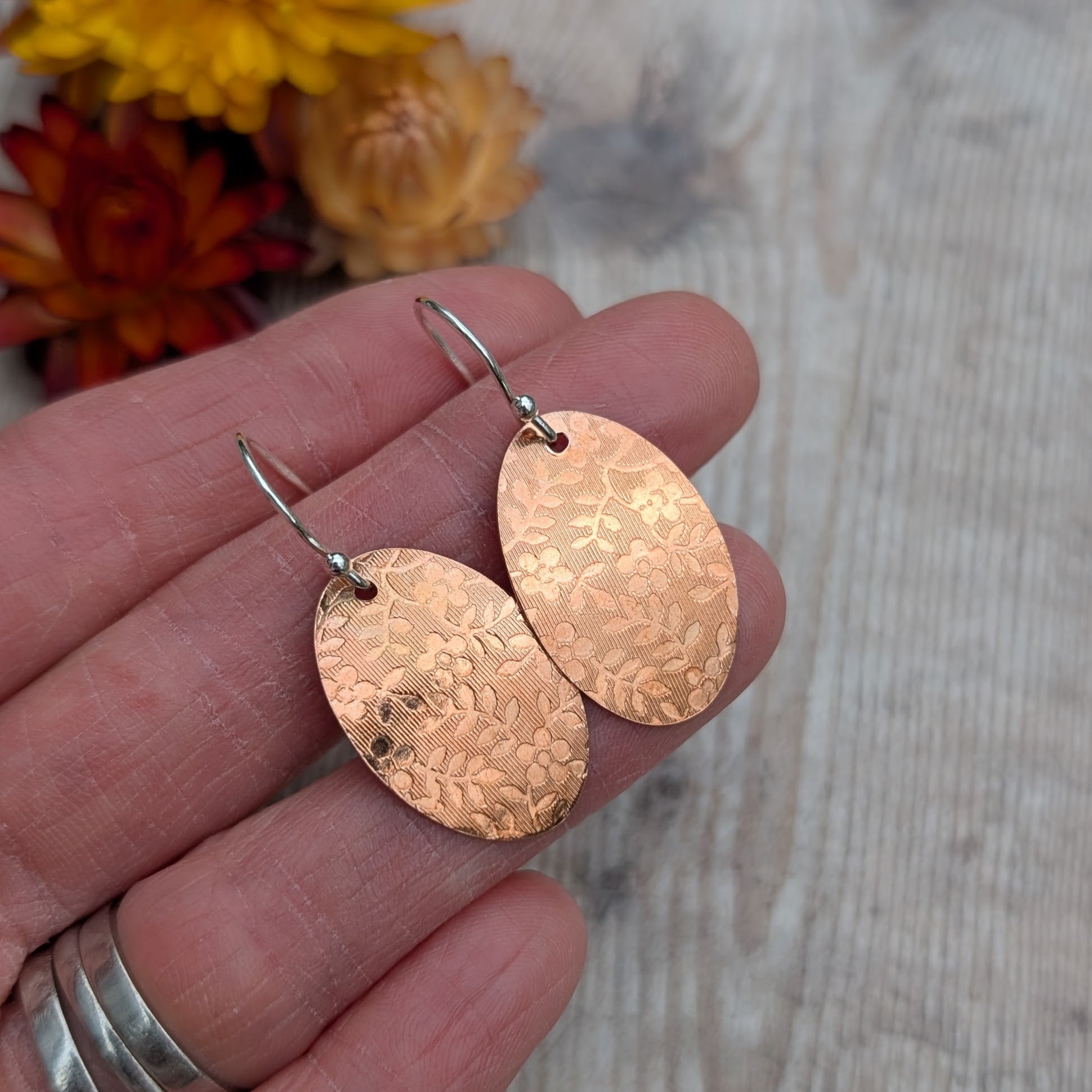 Copper floral pattern oval disc earrings held in a hand to show size and detail.