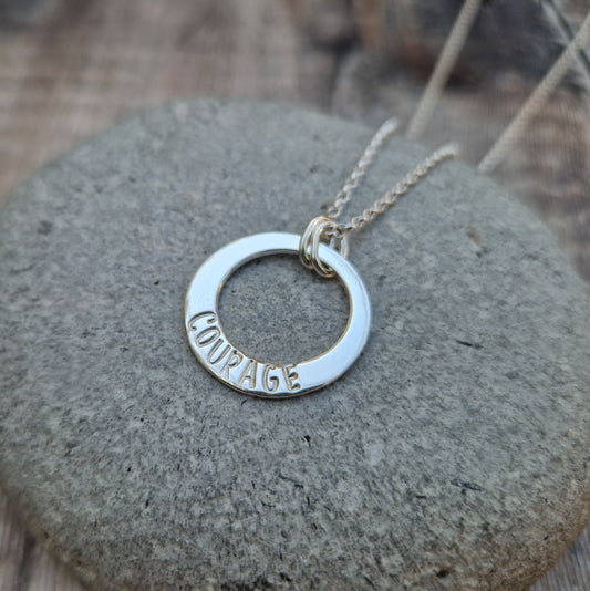 Sterling Silver ‘COURAGE’ necklace. Silver disc with offset hole in centre, looks like a thick circle. Attached via two small silver hoops to silver chain. On thicker part of disc, inscription wording hammered into the silver reads COURAGE in capitals. Disc measures approximately 20 mm in diameter. 