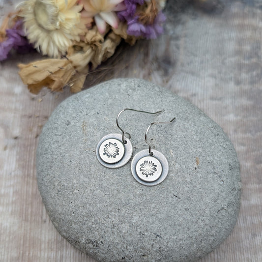 Close-up of oxidised silver earrings with a daisy pattern on each disc, displayed on a smooth stone with dried flowers in the background.