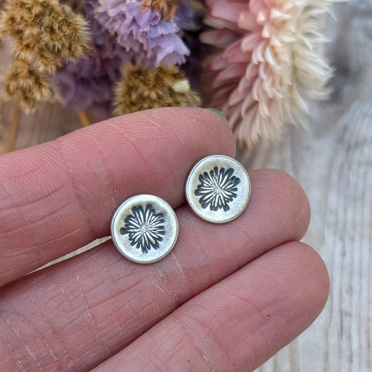 A pair of silver stud earrings with an oxidised daisy pattern on circular discs, held between fingers. Dried flowers in soft purple and beige tones are blurred in the background.