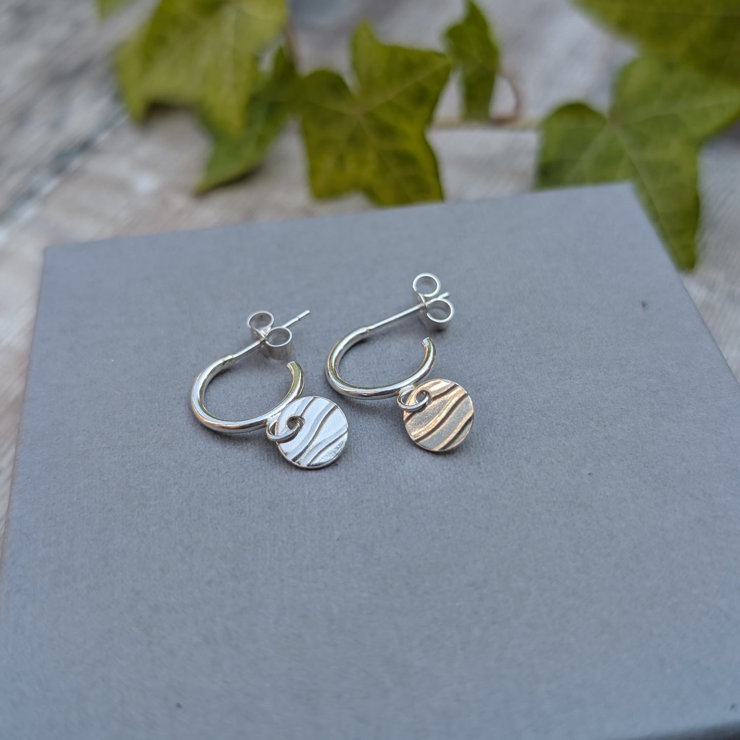 Silver disc drop hoop earrings handmade from recycled Sterling Silver, featuring hand stamped wave pattern discs hanging from small silver hoops on a grey display box.