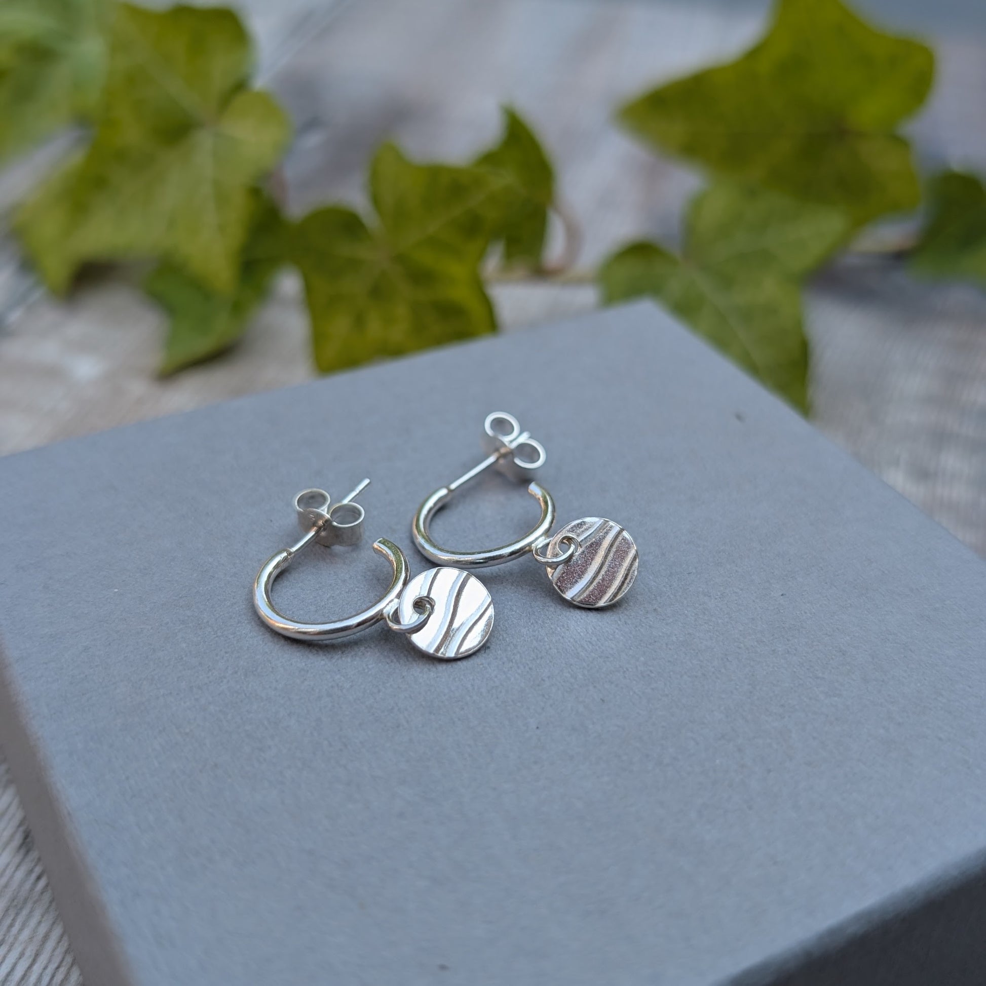 Silver disc drop hoop earrings handmade from recycled Sterling Silver, displayed on a grey jewellery box with green ivy in the background.