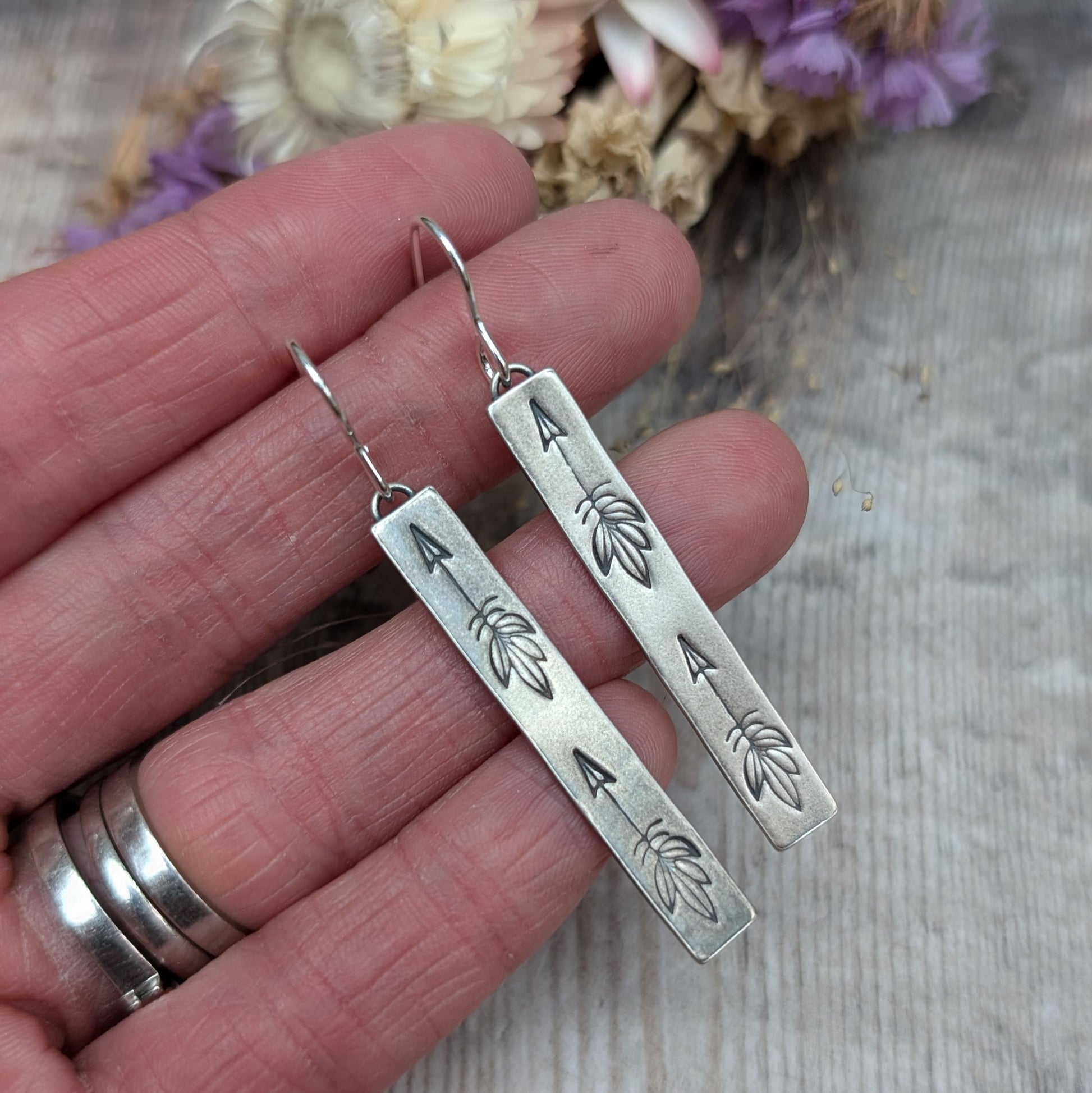 Each earring is an Oxidised Sterling Silver rectangular shaped pendant suspended from a silver earring wire. Stamped on the rectangular shape are horizonal arrows with feather tails, two per earring. The oxidisation has made parts of the pattern darker and lighter in places.