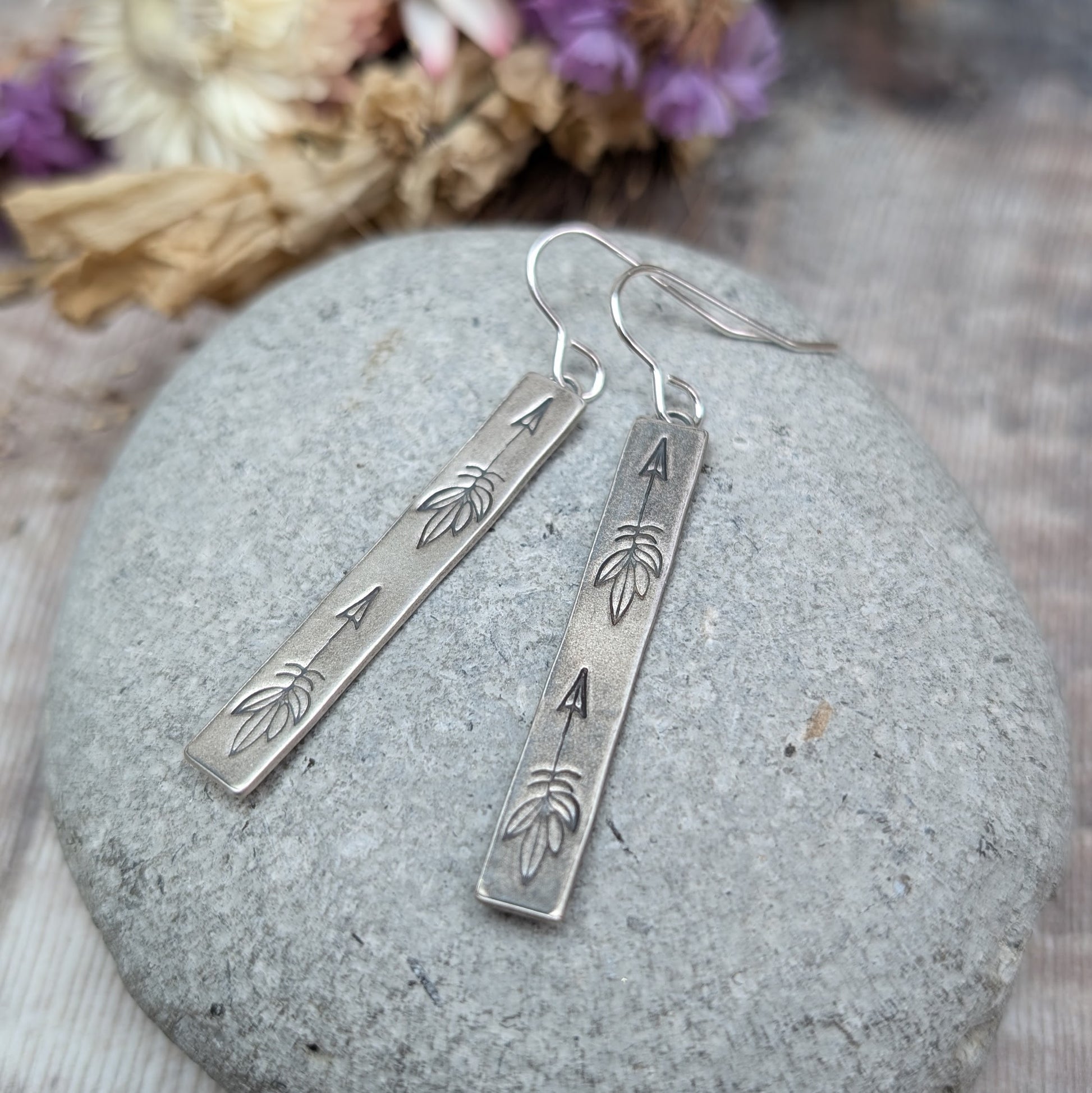 Each earring is an Oxidised Sterling Silver rectangular shaped pendant suspended from a silver earring wire. Stamped on the rectangular shape are horizonal arrows with feather tails, two per earring. The oxidisation has made parts of the pattern darker and lighter in places.