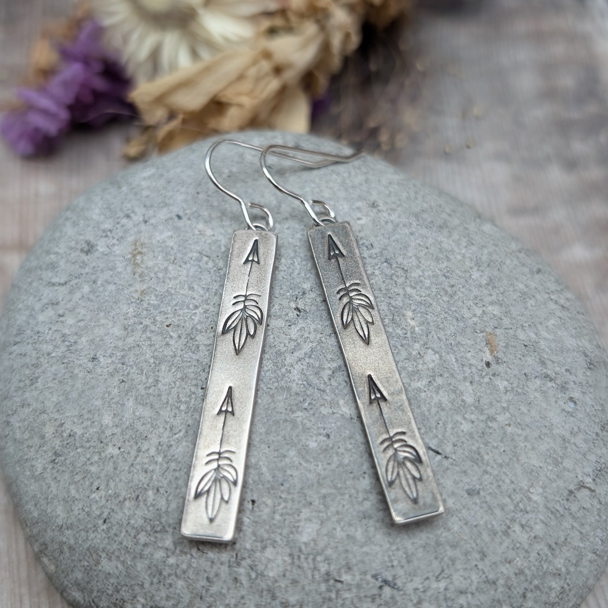 Each earring is an Oxidised Sterling Silver rectangular shaped pendant suspended from a silver earring wire. Stamped on the rectangular shape are horizonal arrows with feather tails, two per earring. The oxidisation has made parts of the pattern darker and lighter in places.