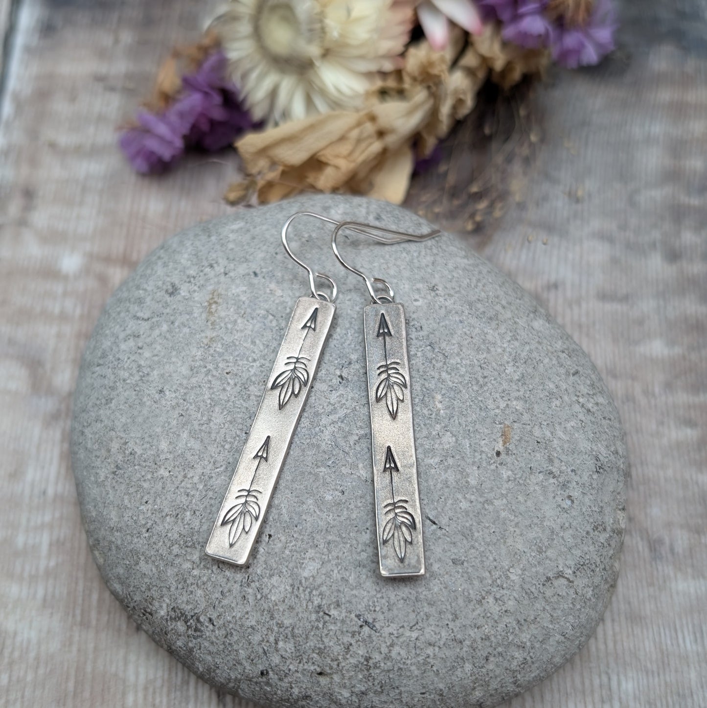 Each earring is an Oxidised Sterling Silver rectangular shaped pendant suspended from a silver earring wire. Stamped on the rectangular shape are horizonal arrows with feather tails, two per earring. The oxidisation has made parts of the pattern darker and lighter in places.