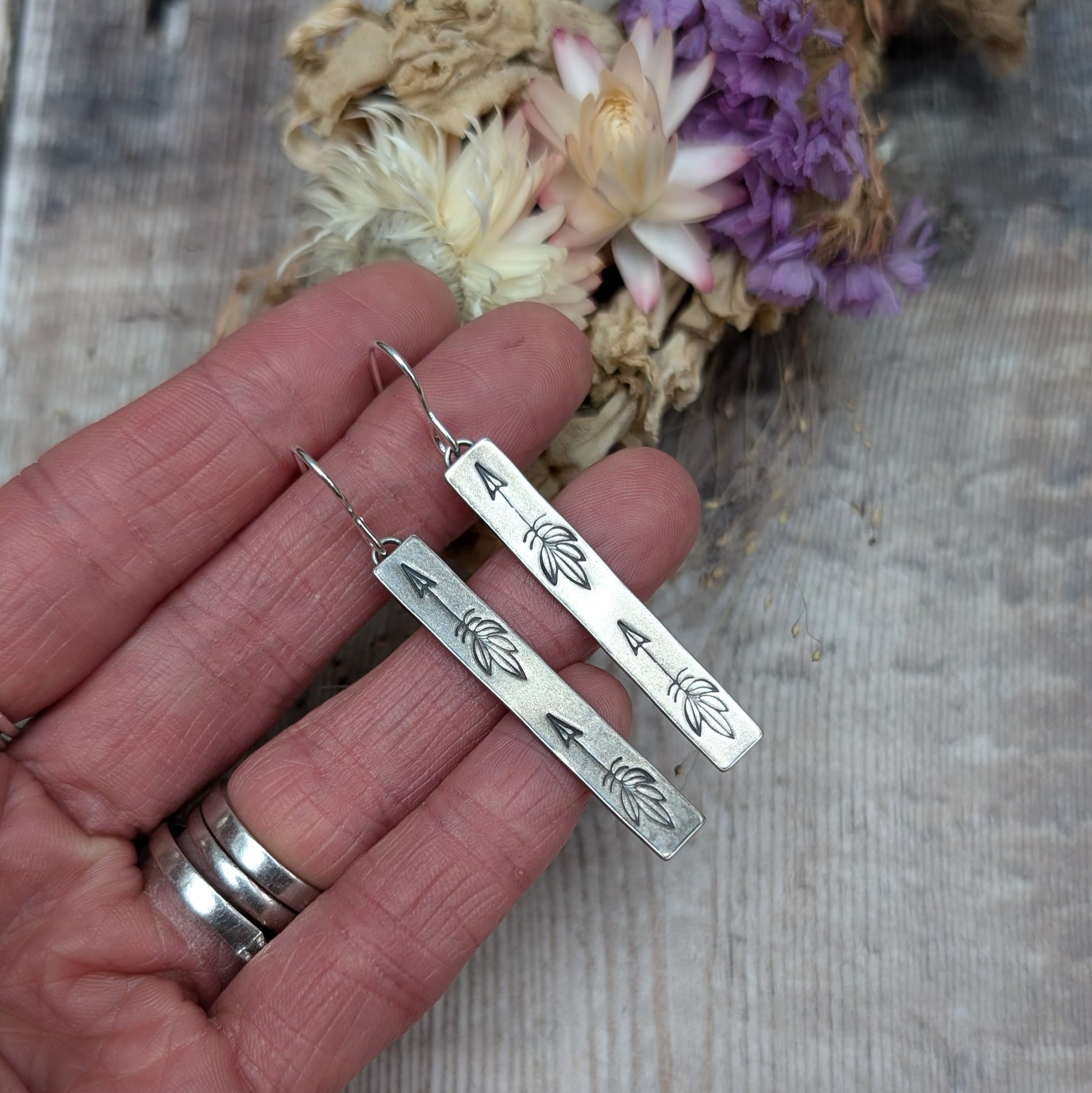 Each earring is an Oxidised Sterling Silver rectangular shaped pendant suspended from a silver earring wire. Stamped on the rectangular shape are horizonal arrows with feather tails, two per earring. The oxidisation has made parts of the pattern darker and lighter in places.