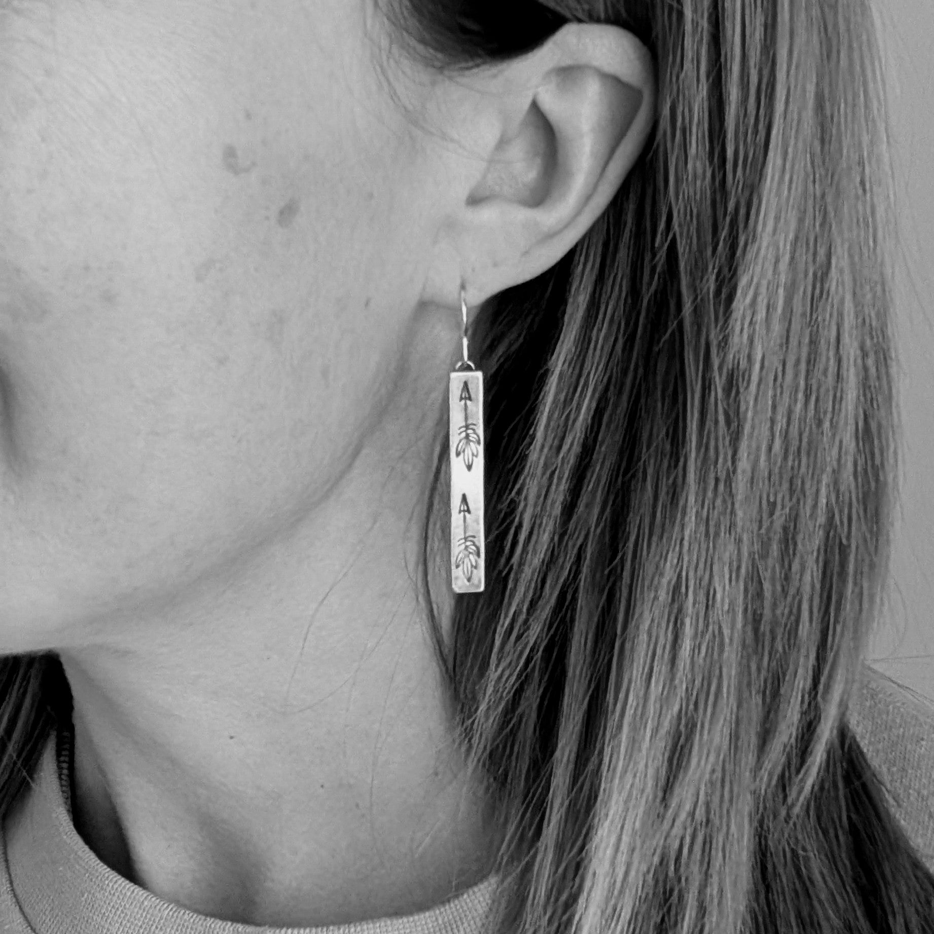 Each earring is an Oxidised Sterling Silver rectangular shaped pendant suspended from a silver earring wire. Stamped on the rectangular shape are horizonal arrows with feather tails, two per earring. The oxidisation has made parts of the pattern darker and lighter in places.