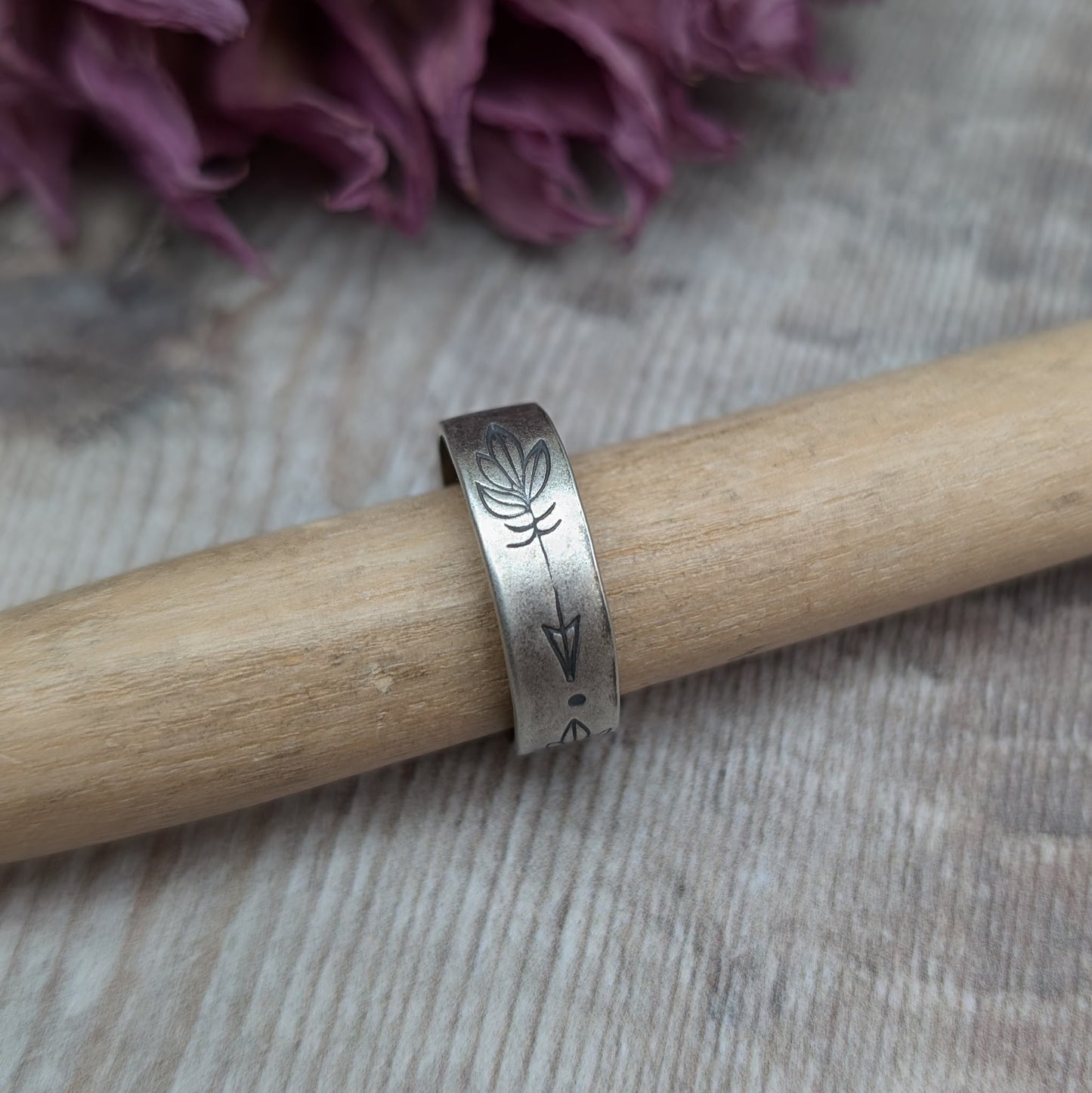 A close-up of a handmade silver ring with a 5mm wide band, featuring hand-stamped feather designs. The ring has an oxidized finish and is displayed on a wooden rod, with dried flowers in the background.