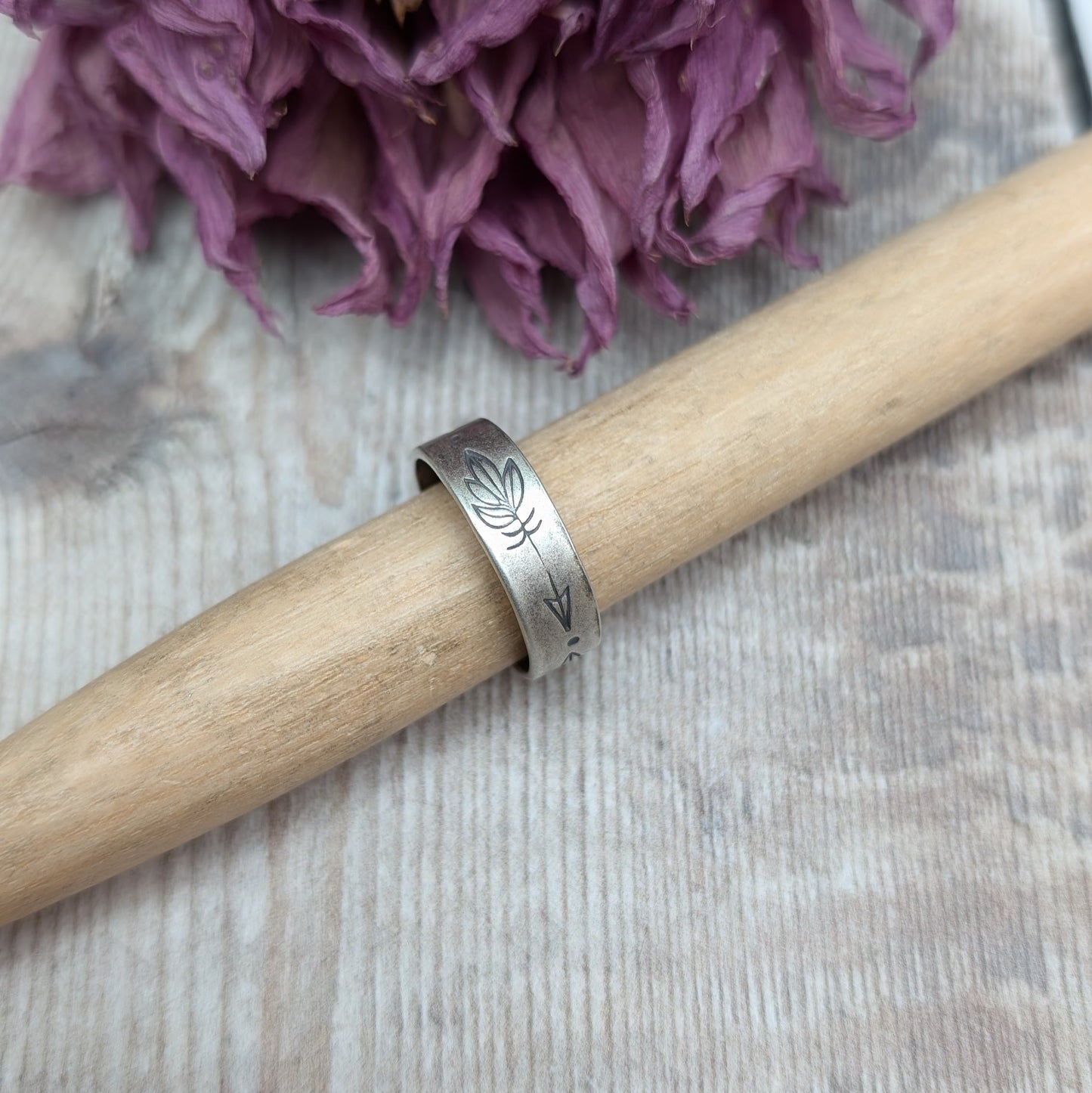 A close-up of a handmade silver ring with a 5mm wide band, featuring hand-stamped feather designs. The ring has an oxidized finish and is displayed on a wooden rod, with dried flowers in the background.