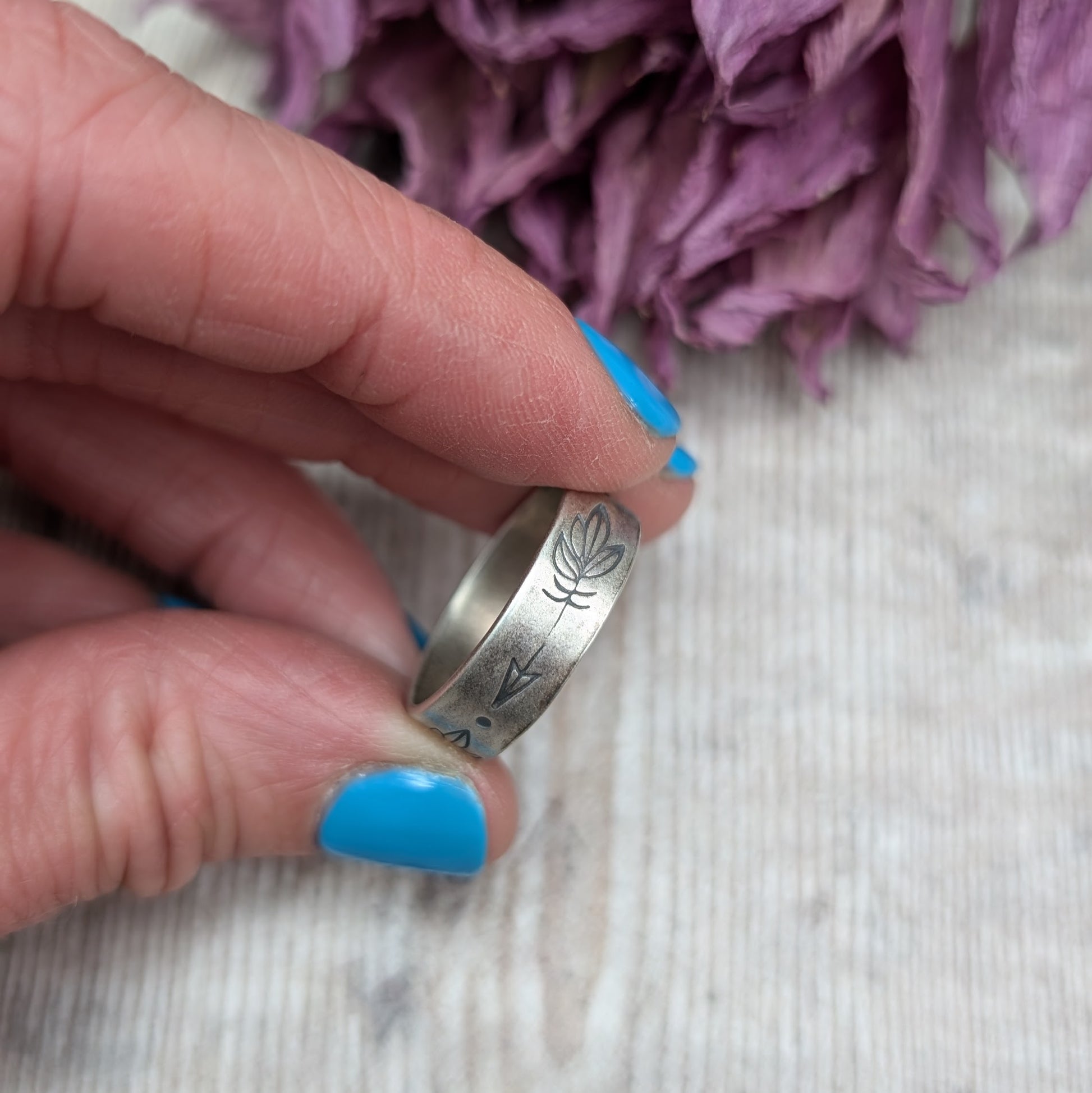 A close-up of a hand holding a handmade silver ring with a 5mm wide band, featuring hand-stamped feather designs. The ring has an oxidized finish and dried purple flowers are in the background.