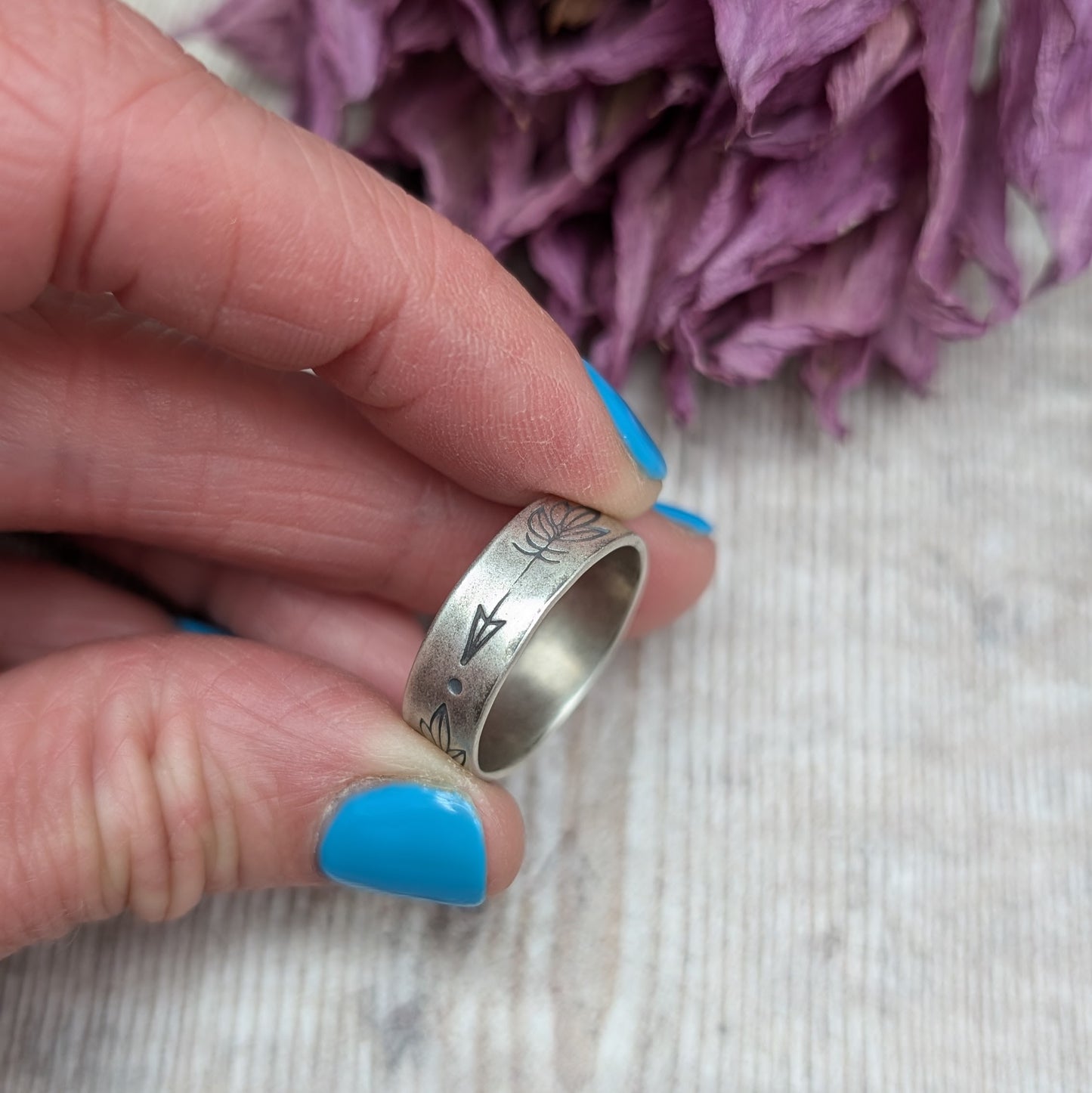 A close-up of a hand holding a handmade silver ring with a 5mm wide band, featuring hand-stamped feather designs. The ring has an oxidized finish and dried purple flowers are in the background.