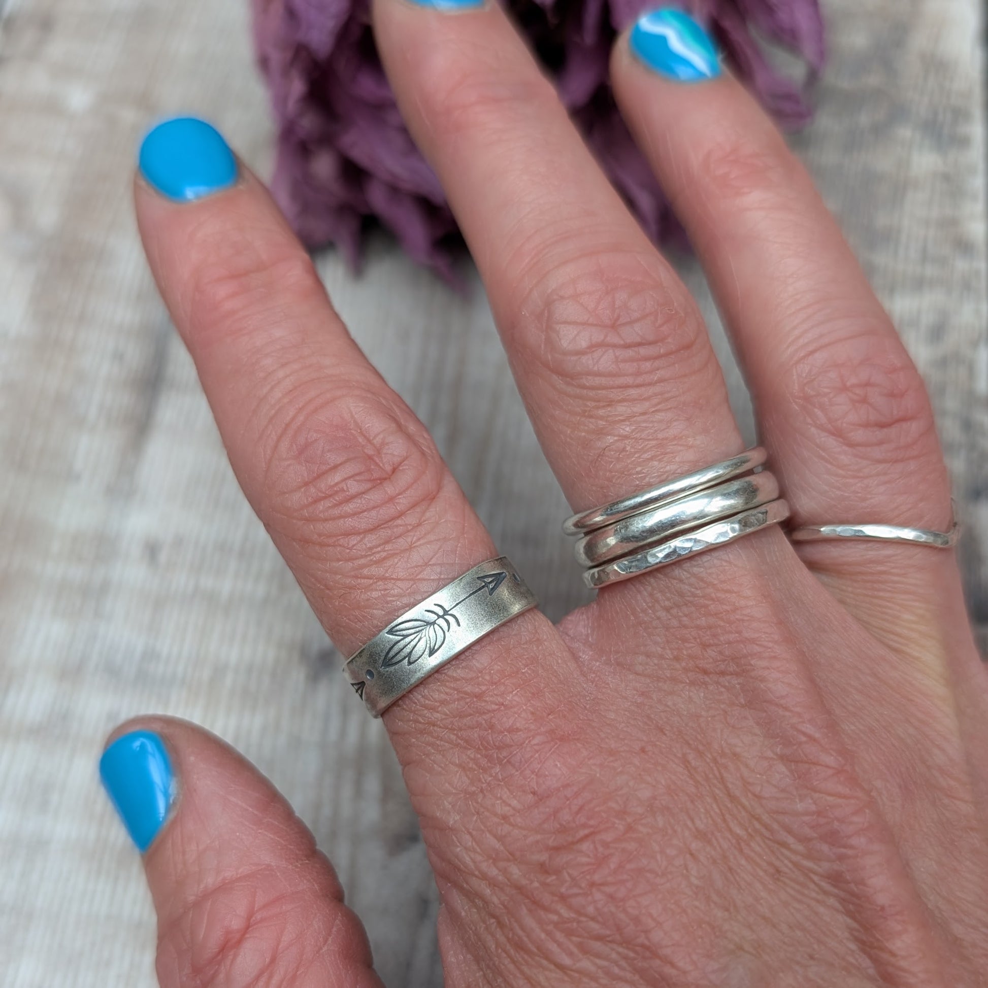 A hand showcasing a handmade silver ring with a 5mm wide band and hand-stamped feather designs. The ring has an oxidized finish.