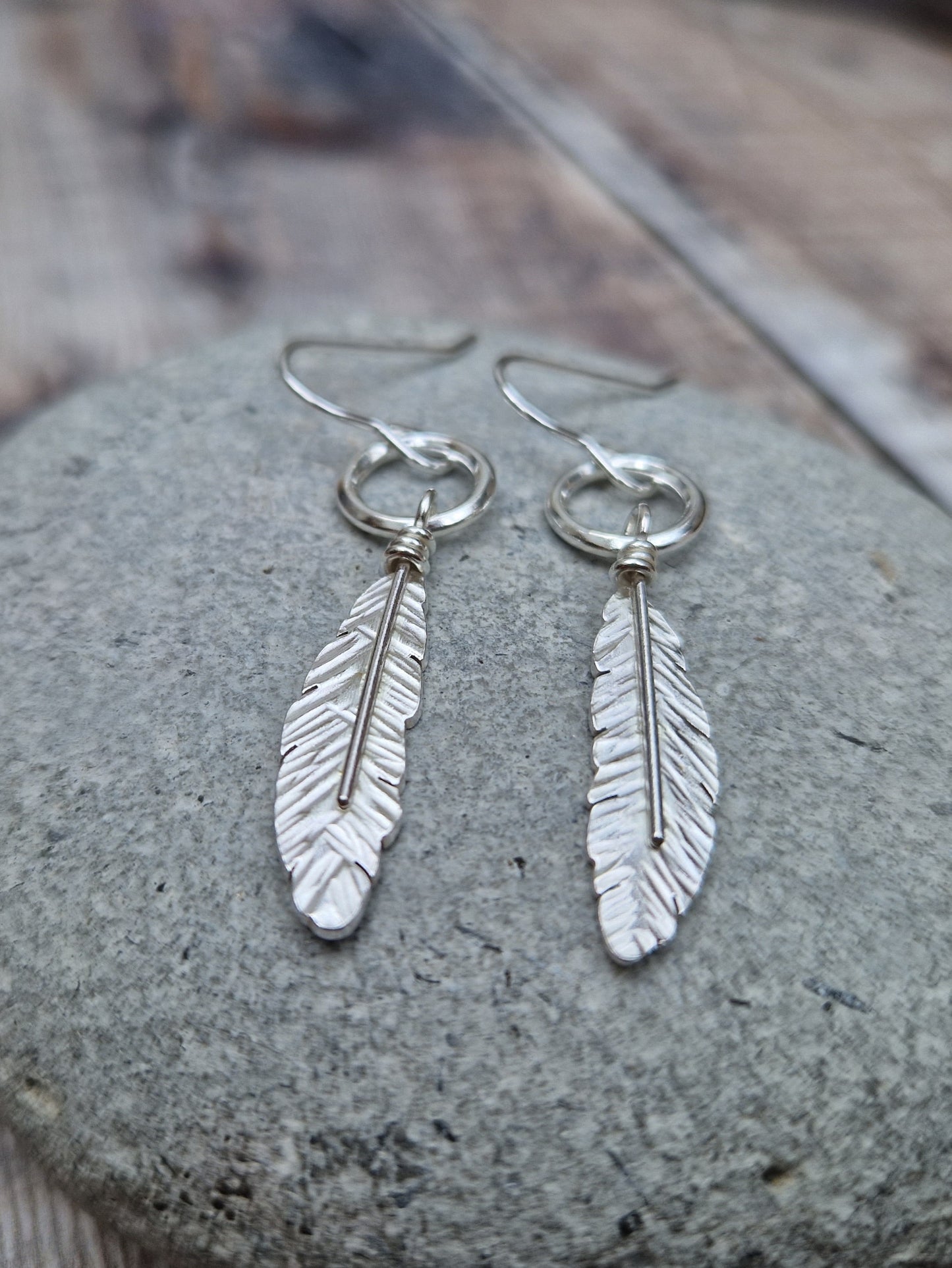 Silver dangle earrings featuring a detailed feather design suspended from a small circular hoop. The earrings rest on a smooth stone, highlighting the intricate texture of the feathers against the natural backdrop.