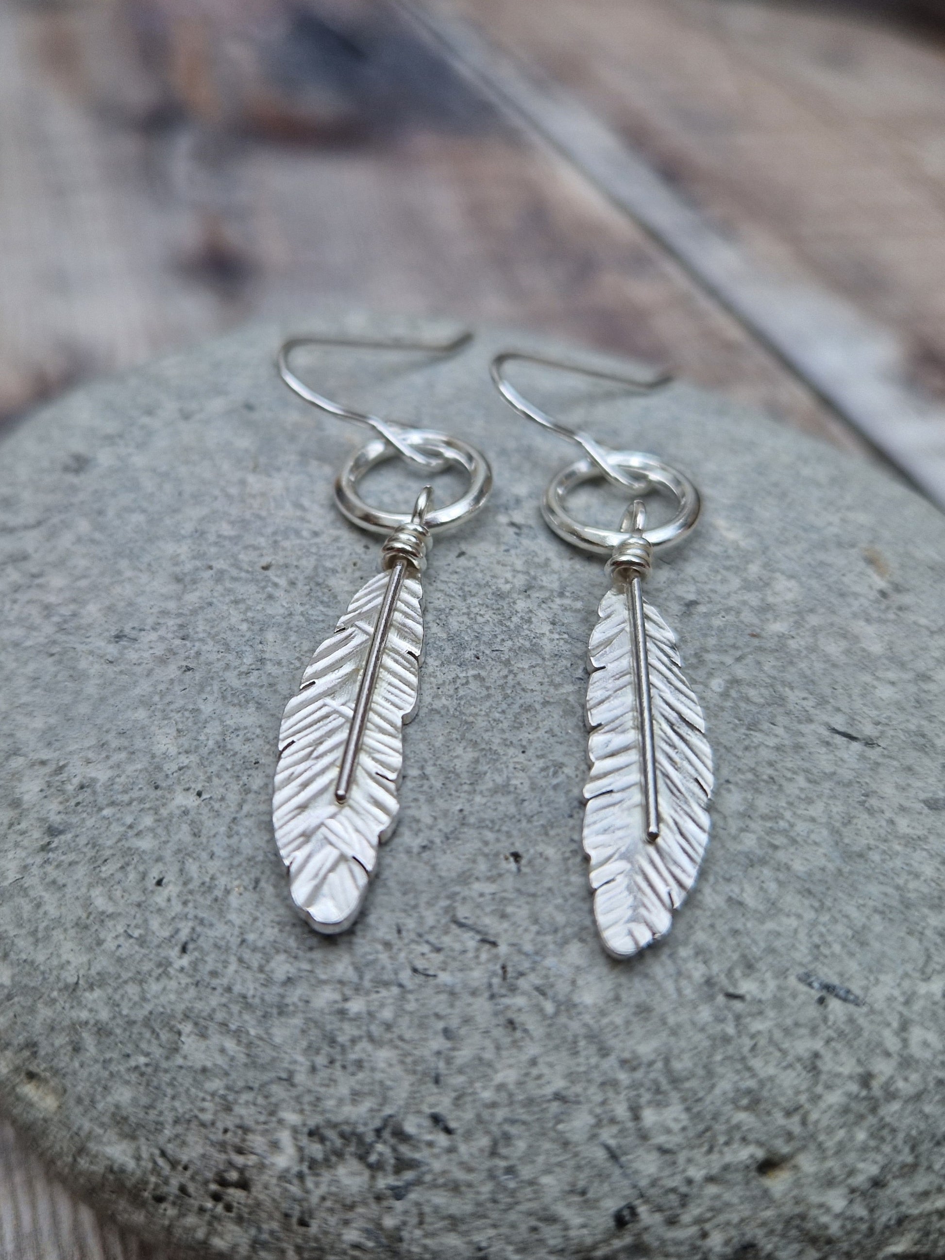 Silver dangle earrings featuring a detailed feather design suspended from a small circular hoop. The earrings rest on a smooth stone, highlighting the intricate texture of the feathers against the natural backdrop.