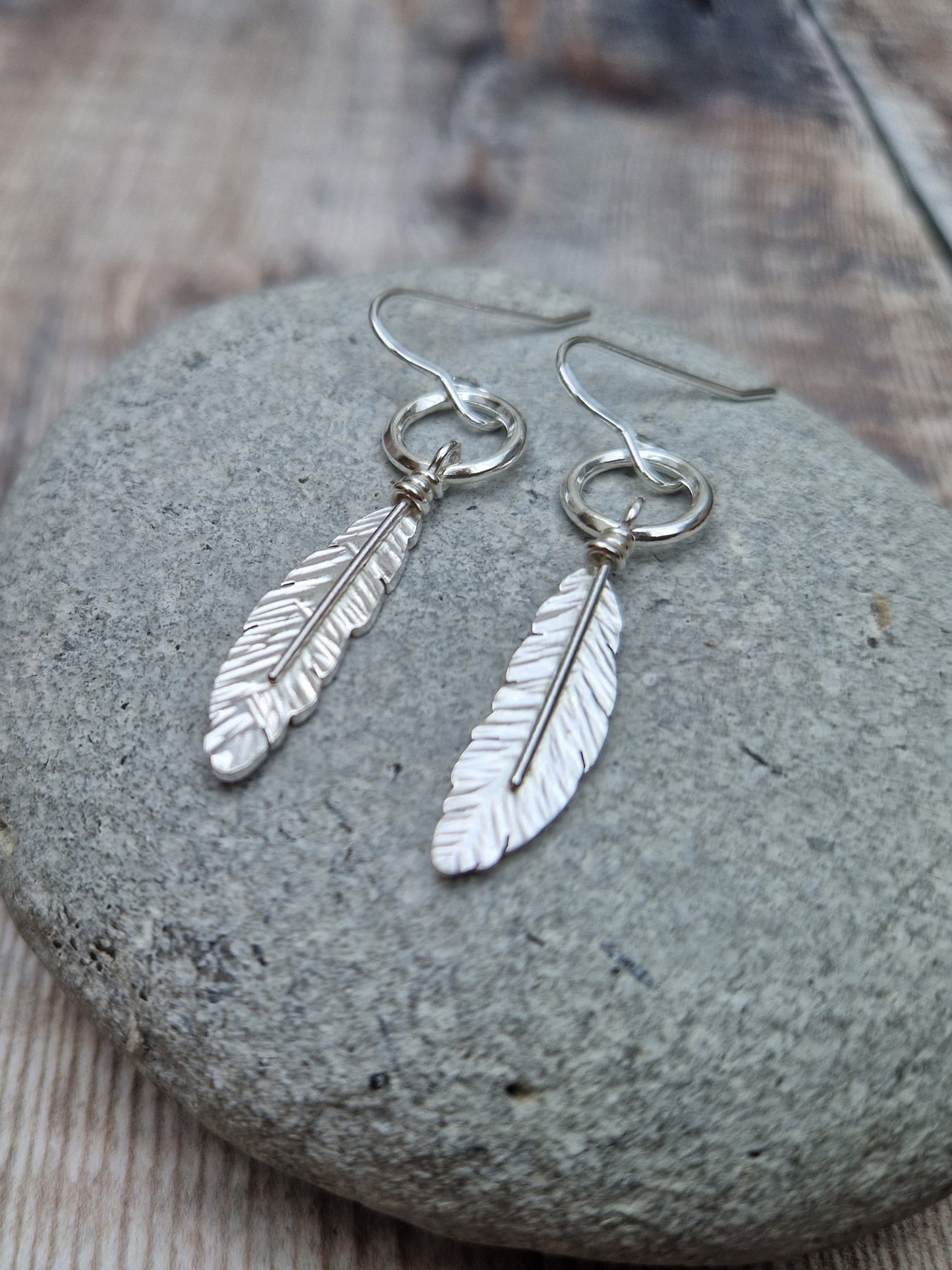 Silver dangle earrings featuring a detailed feather design suspended from a small circular hoop. The earrings rest on a smooth stone, highlighting the intricate texture of the feathers against the natural backdrop.