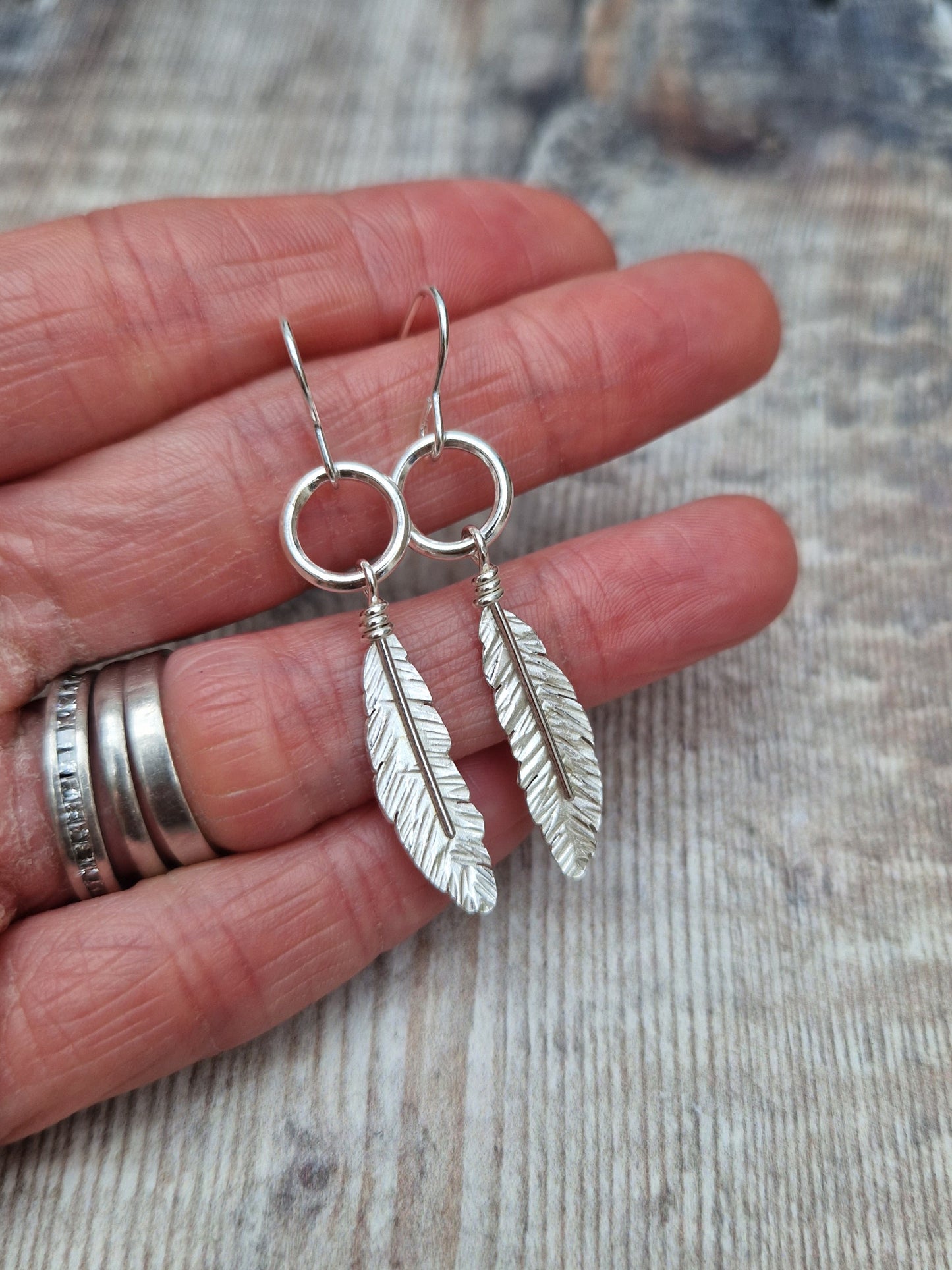 Hand holding a pair of silver dangle earrings with feather charms hanging from circular hoops. The delicate feather details are showcased against the skin, emphasizing the earrings' lightweight and elegant style.