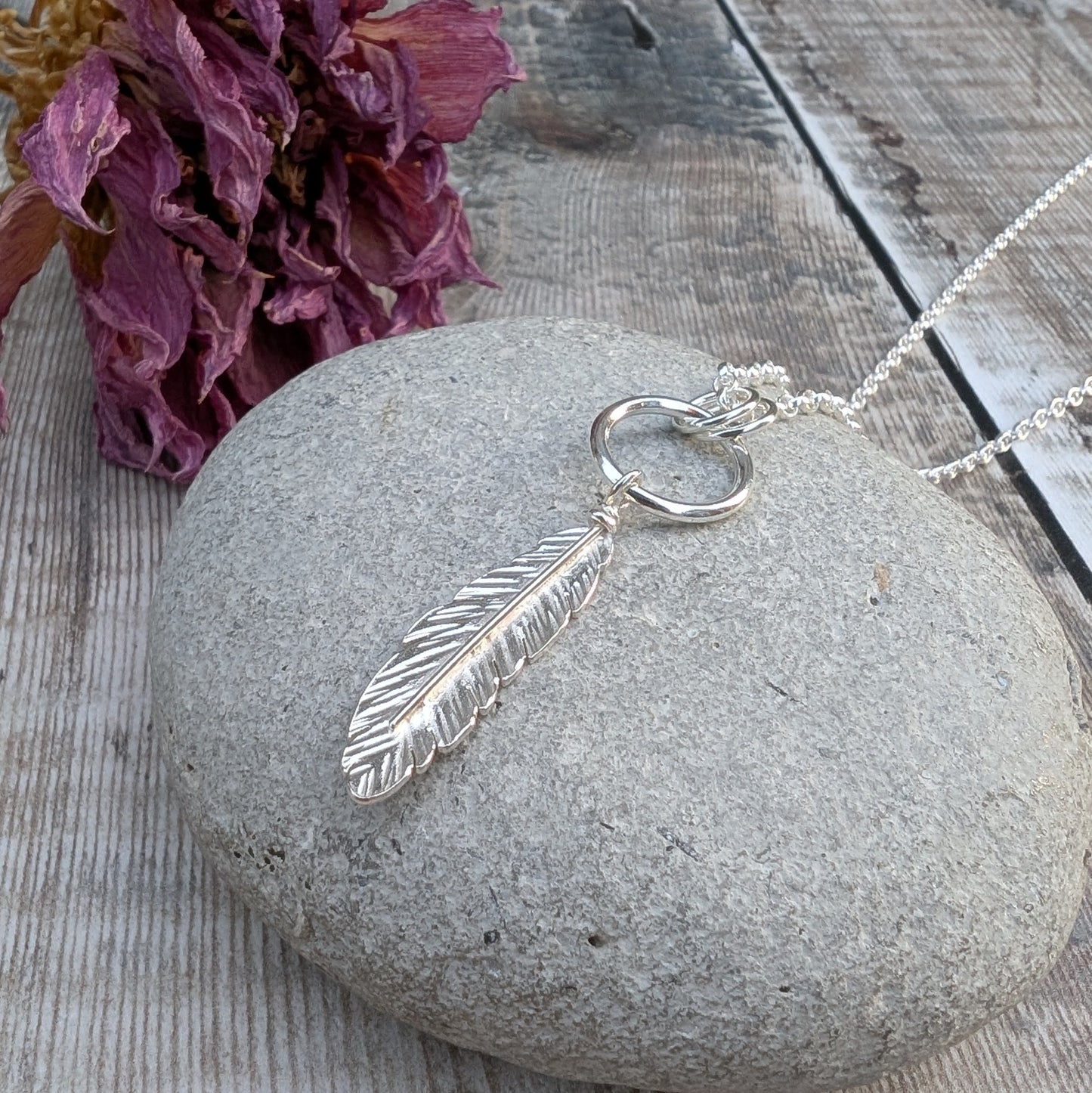 A handmade sterling silver necklace featuring a detailed feather pendant hanging from a circular silver ring, displayed on a smooth grey stone. The necklace chain extends out of the frame, and dried purple flowers are in the background.